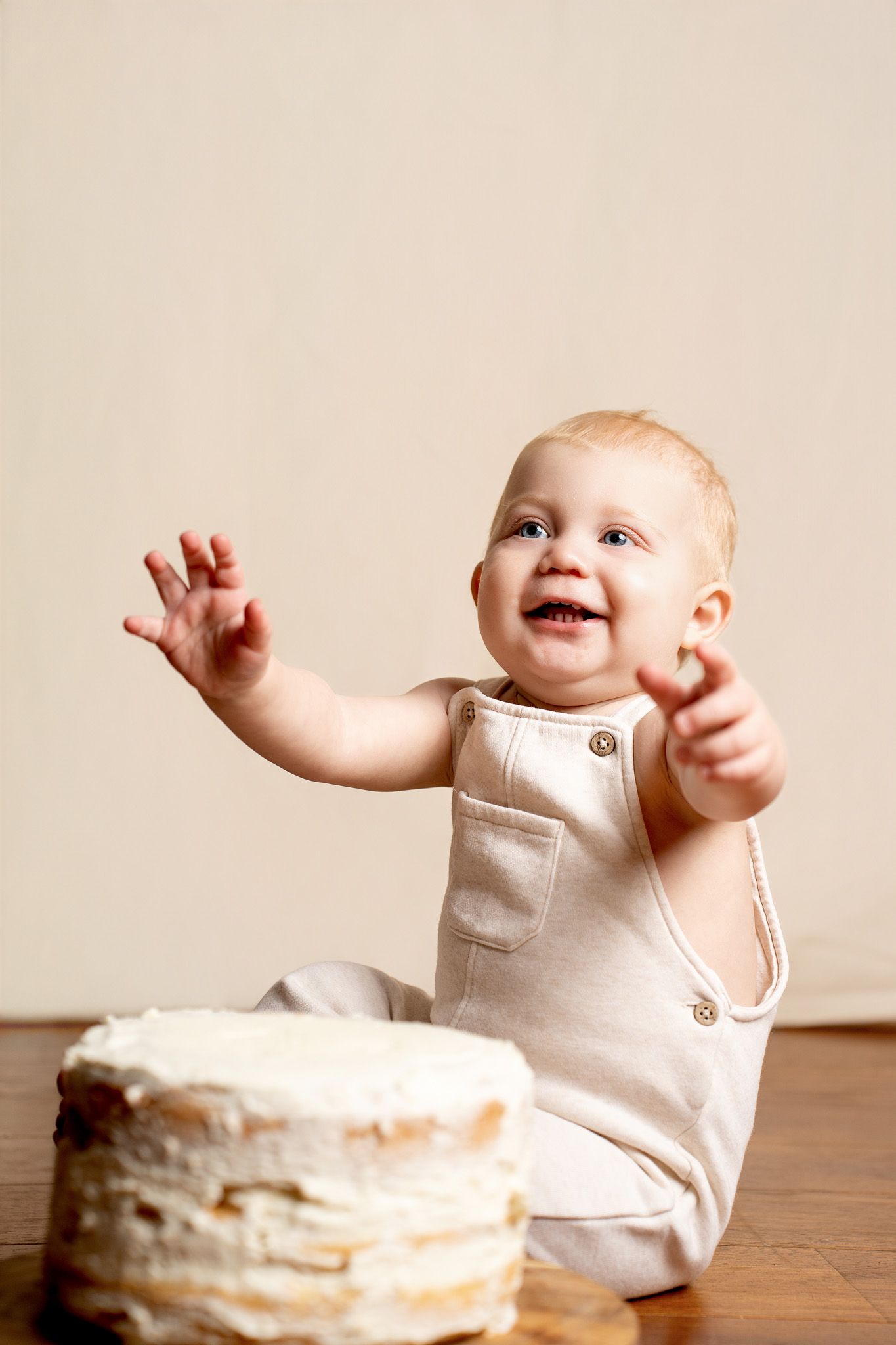 Baby in cream outfit sits next to white birthday cake on wooden surface.