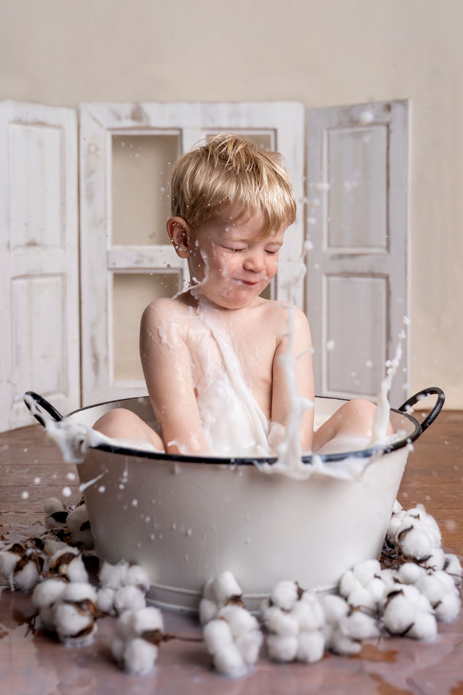 A smiling toddler boy sits in a vintage metal tub splashing milk bath, surrounded by cotton flowers.