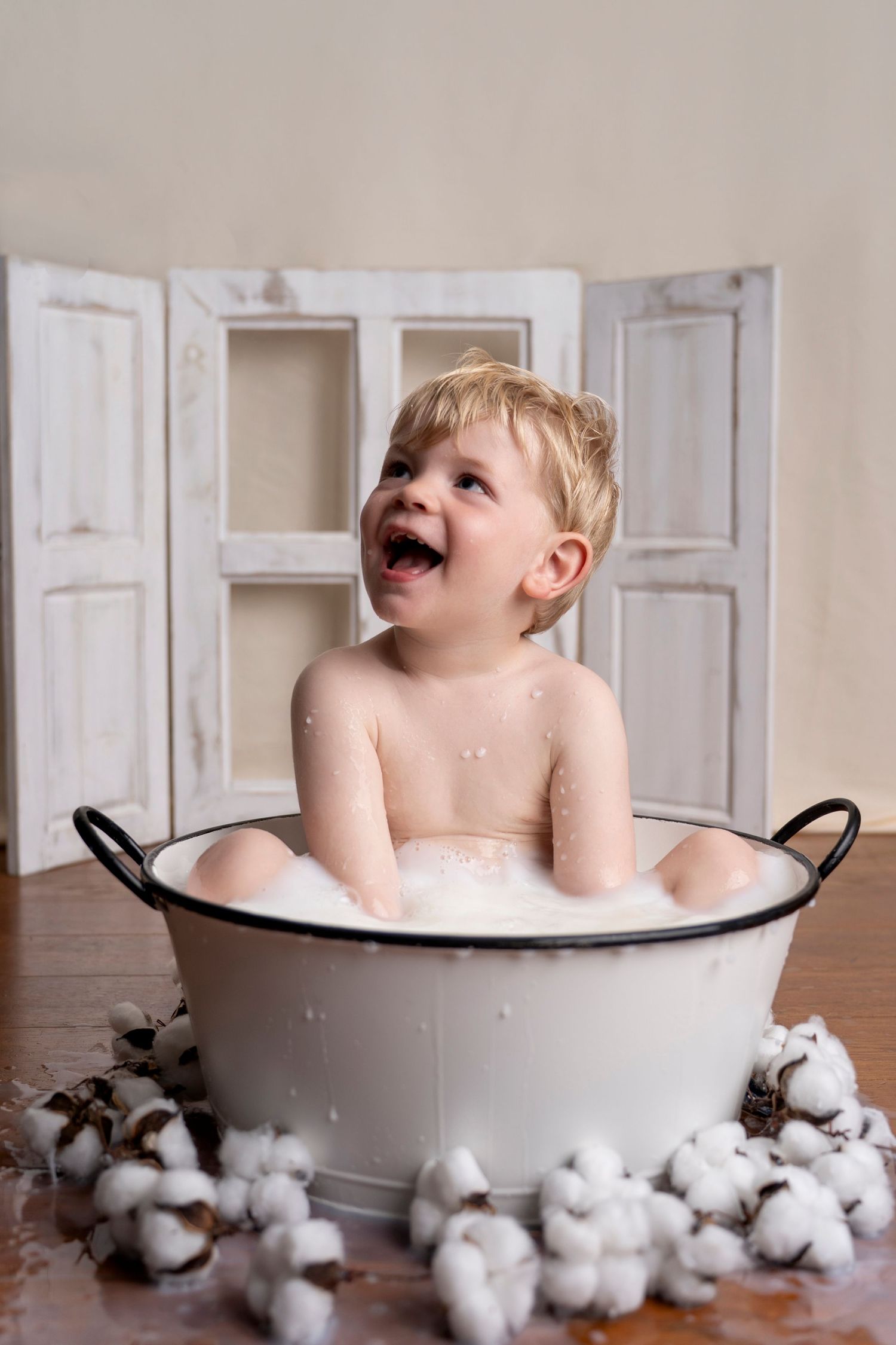 Laughing toddler sitting in a vintage white enamel tub with bubbles, surrounded by cotton flowers on a wooden floor.