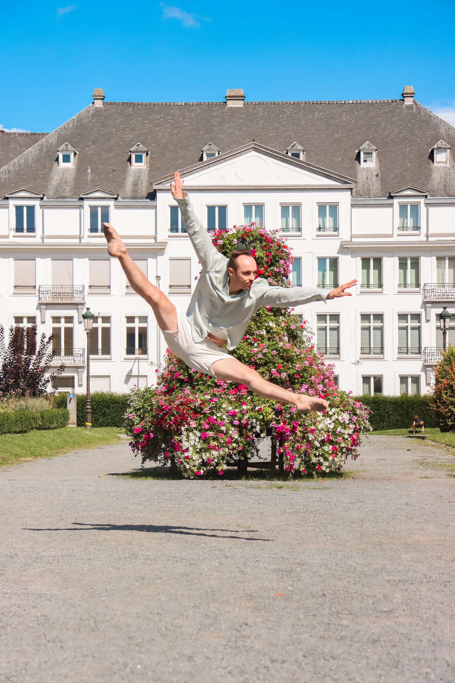 A dancer leaps gracefully in front of a white building with pink flowering bushes.