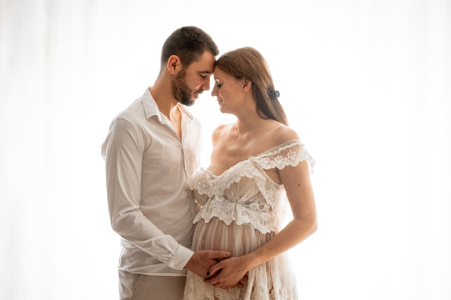 Intimate maternity photoshoot with couple in white lace against bright background.
