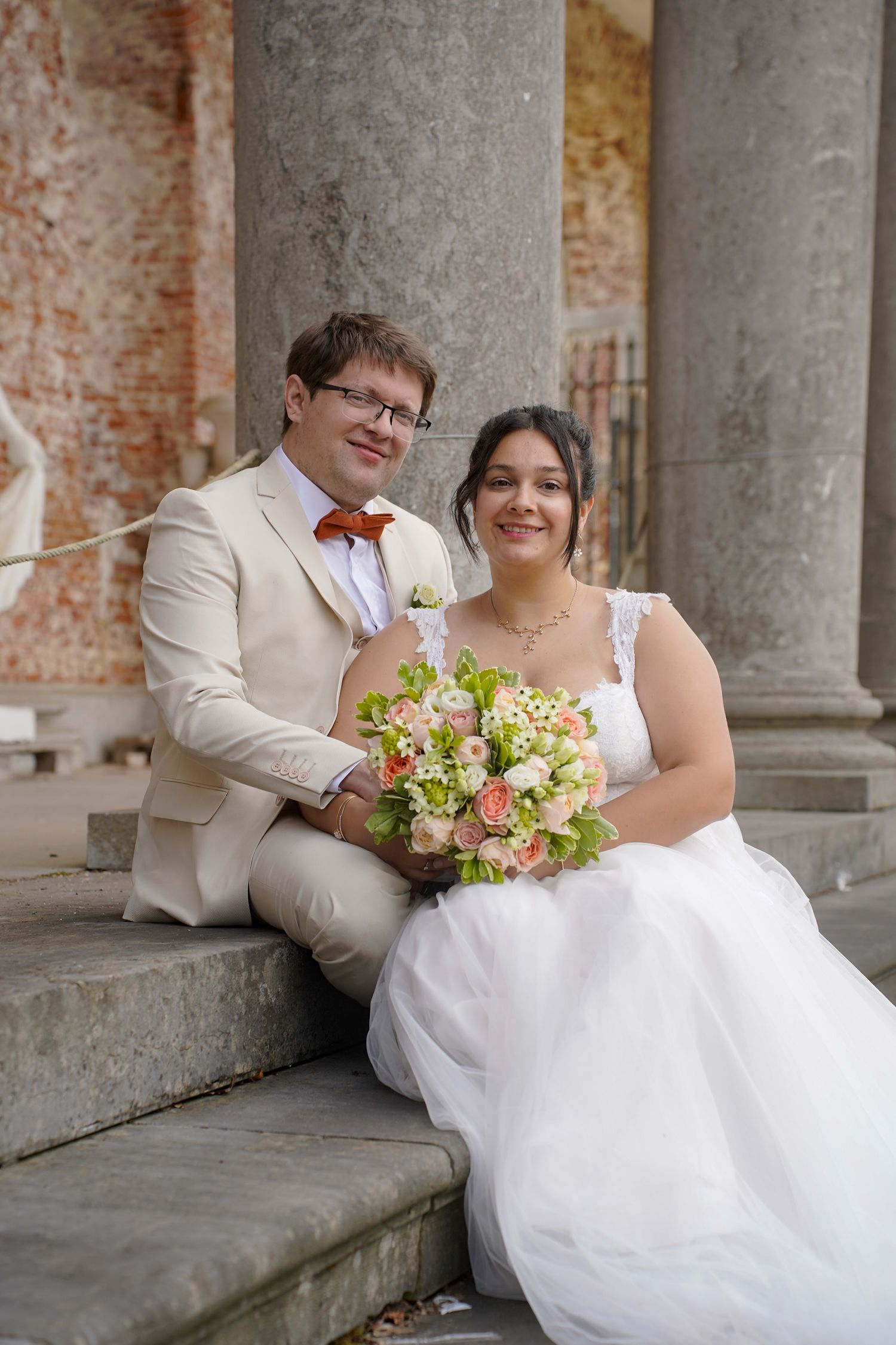 Romantic wedding couple poses together on stone steps near brick columns with floral bouquet.