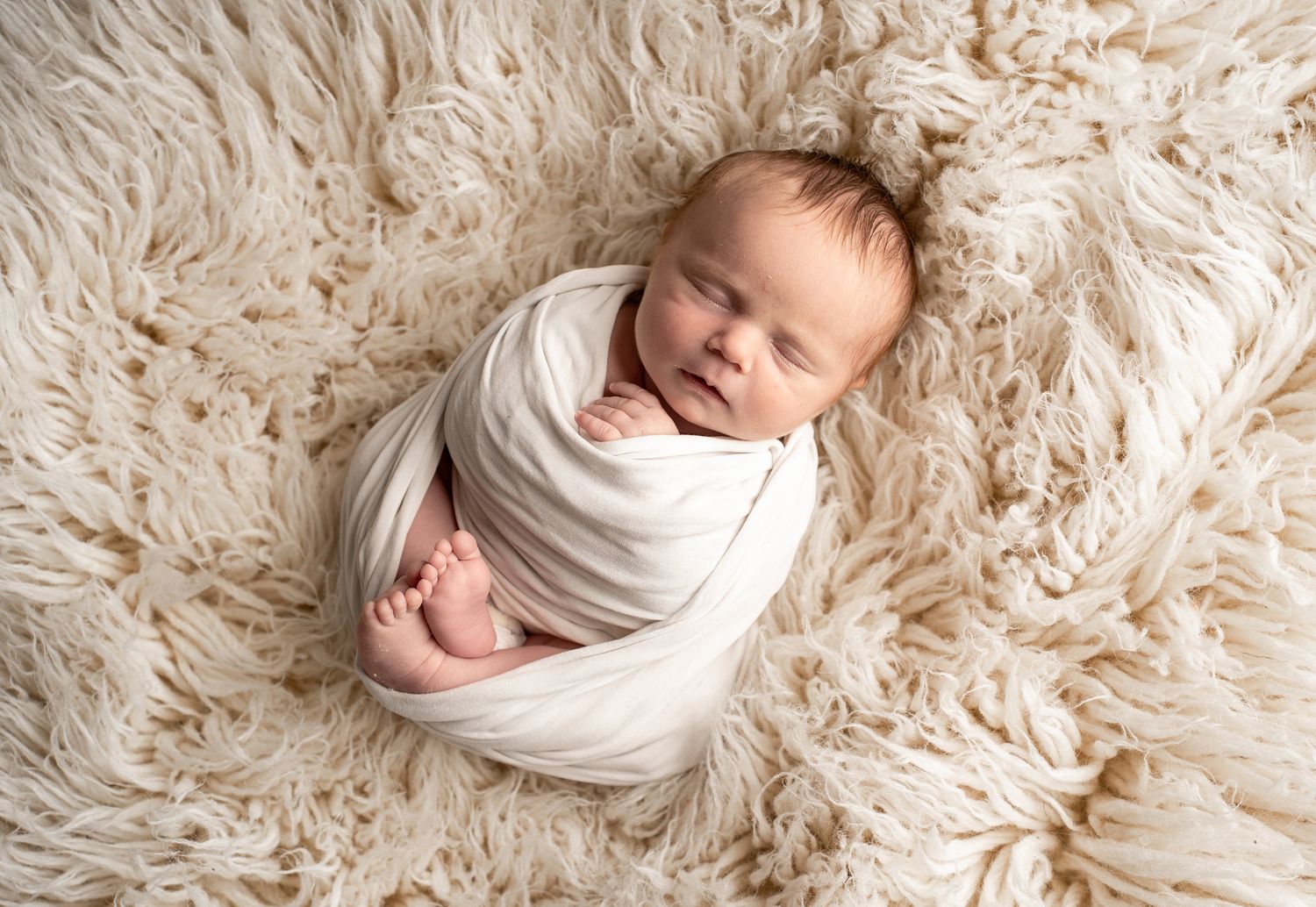 Peaceful newborn baby wrapped in white blanket sleeping on soft furry rug.