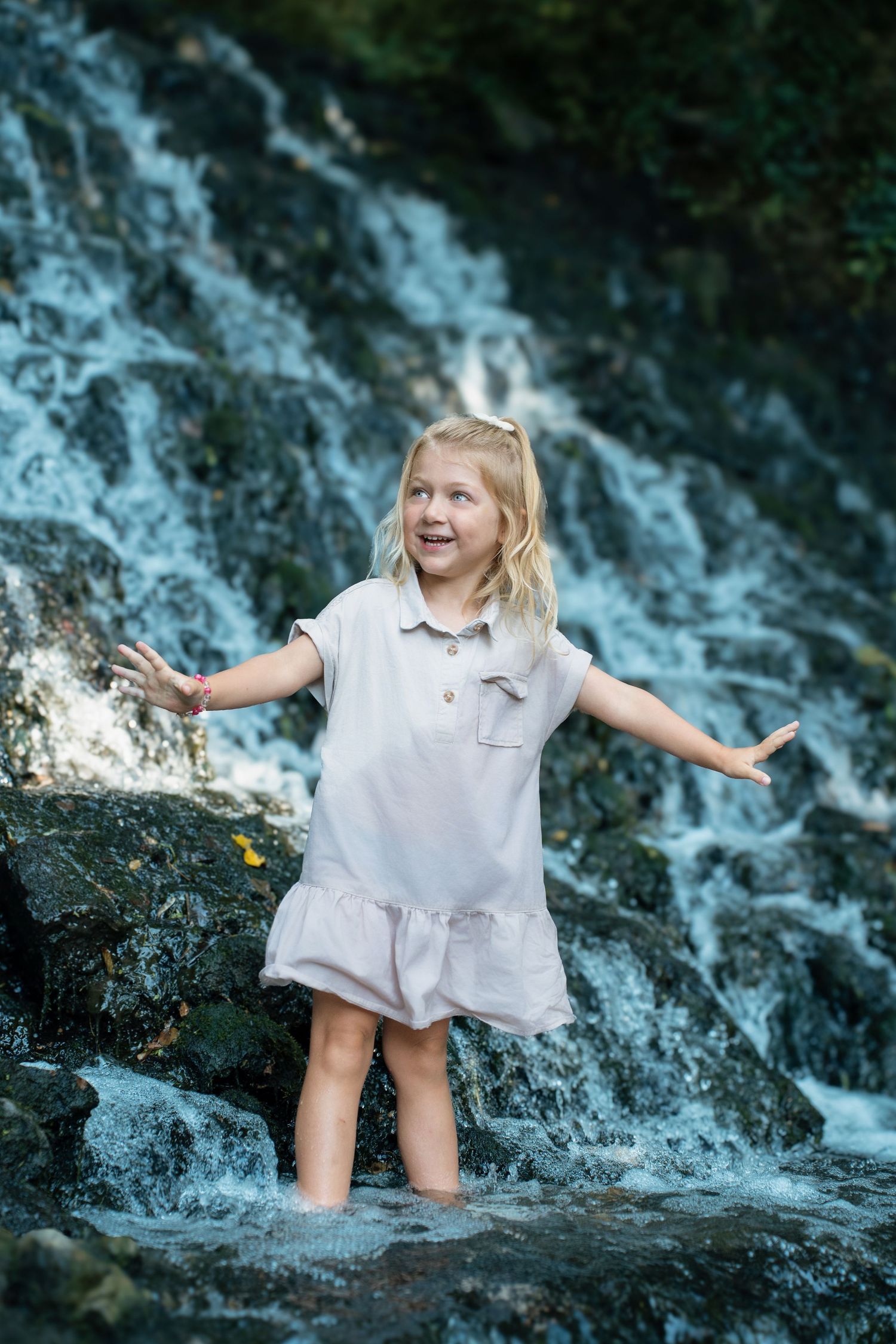 A joyful blonde girl in a white dress stands barefoot in water before a rocky waterfall, arms outstretched.