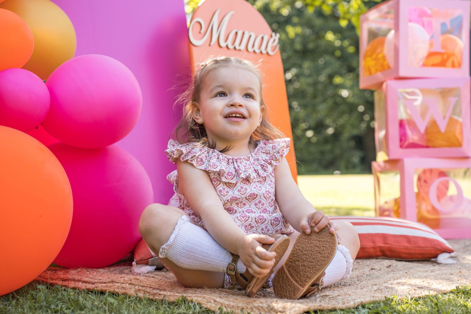 Festive baby photoshoot setup with pink and orange balloons against green outdoor backdrop.