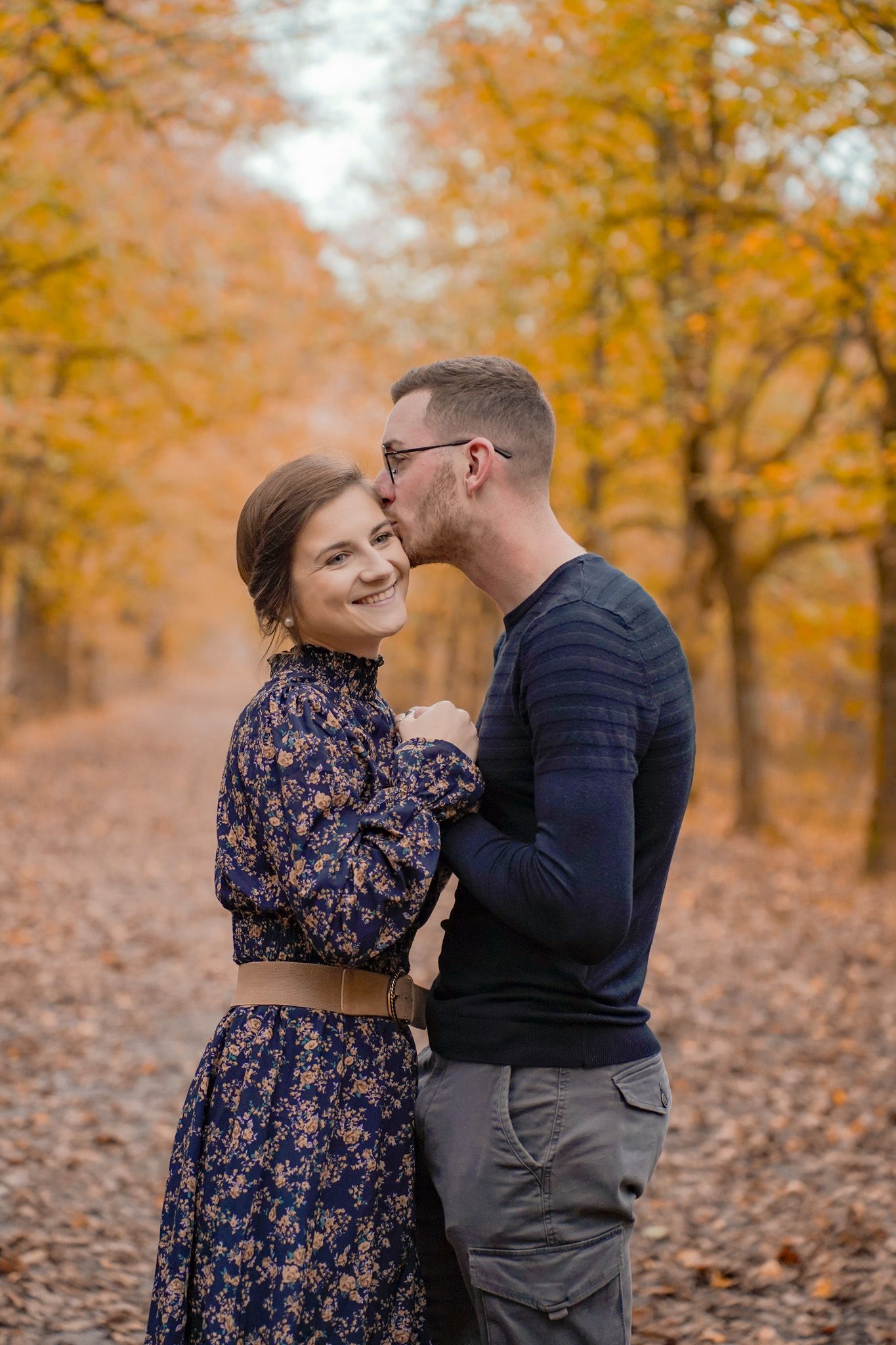 Couple embracing outdoors during autumn with fallen leaves and trees in the background.