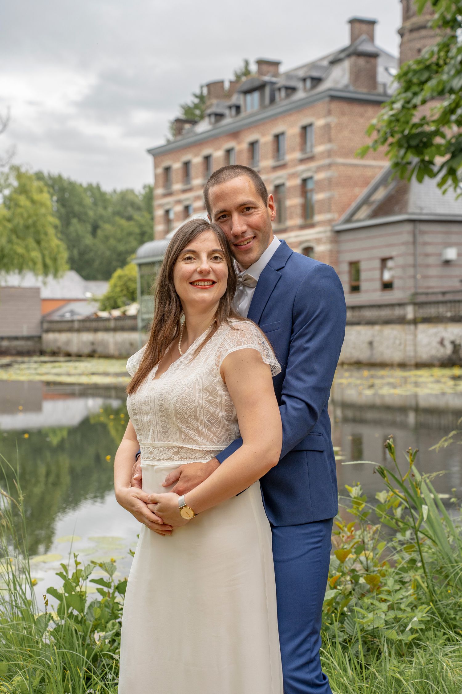A couple poses together by a scenic lake with a historic brick building in the background.