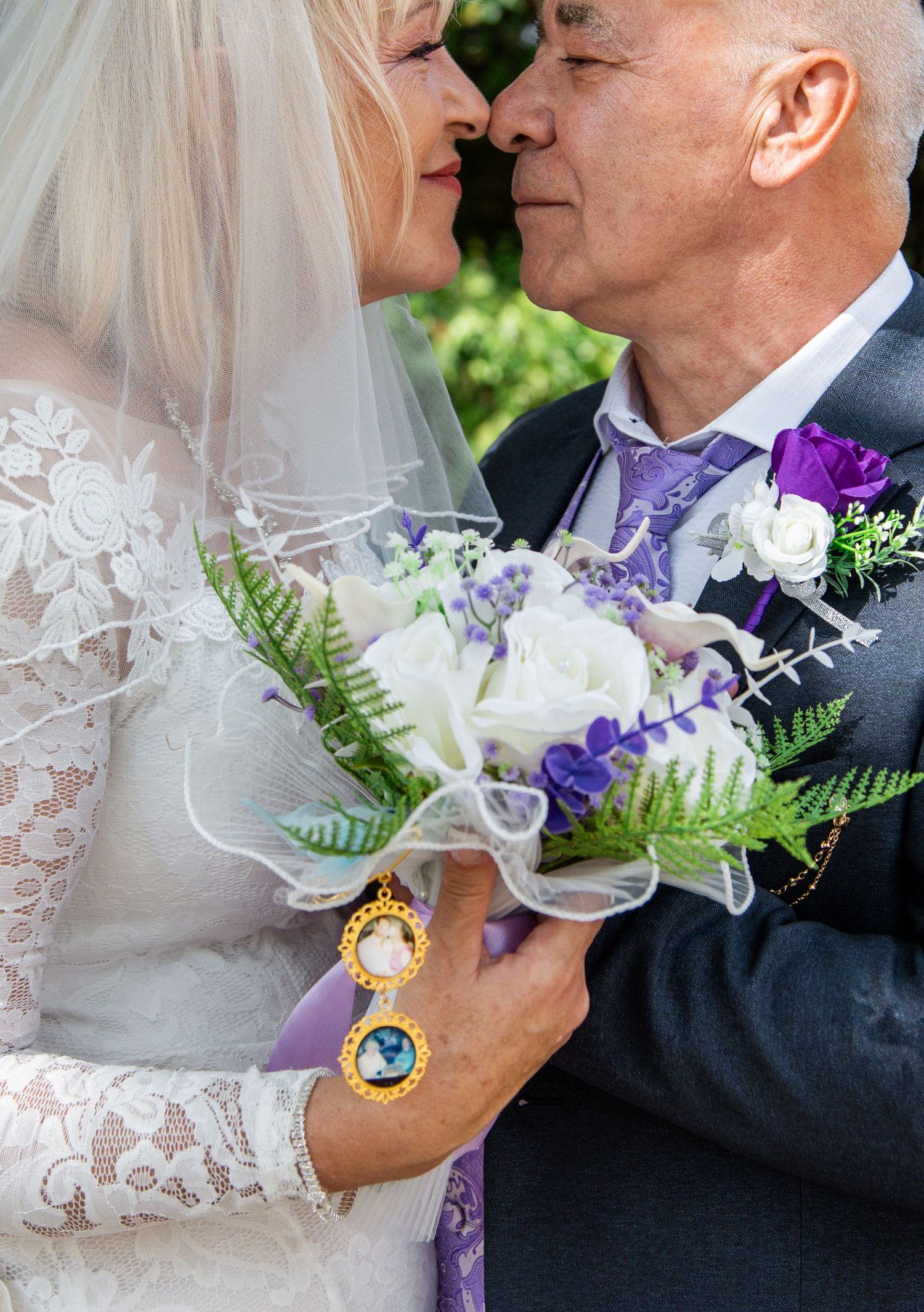 Bride and groom share a romantic moment, holding a white and purple wedding bouquet with fern accents.