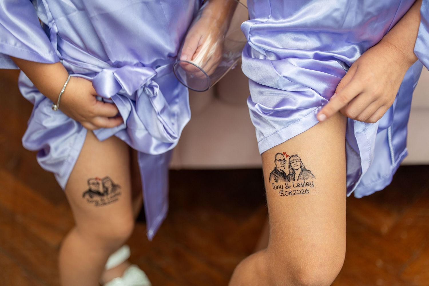 Two bridesmaids in blue satin robes showing matching wedding date tattoos on their legs.