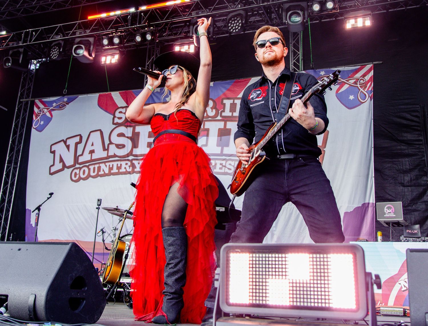 A woman in red dress and man with guitar perform on Nashville Country Music Festival outdoor stage.