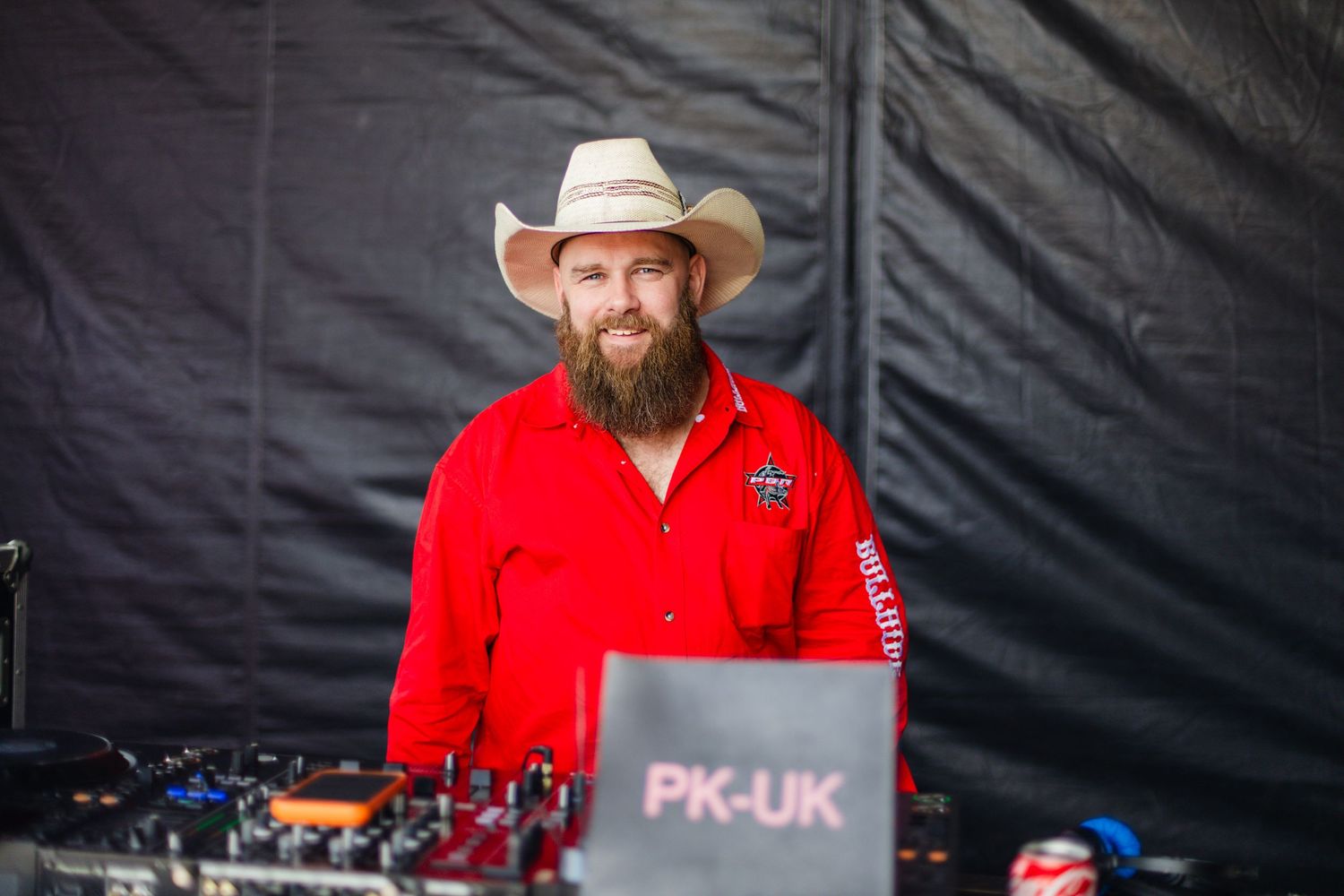 Bearded man in red shirt and cowboy hat DJing at outdoor event behind PK-UK branded equipment.