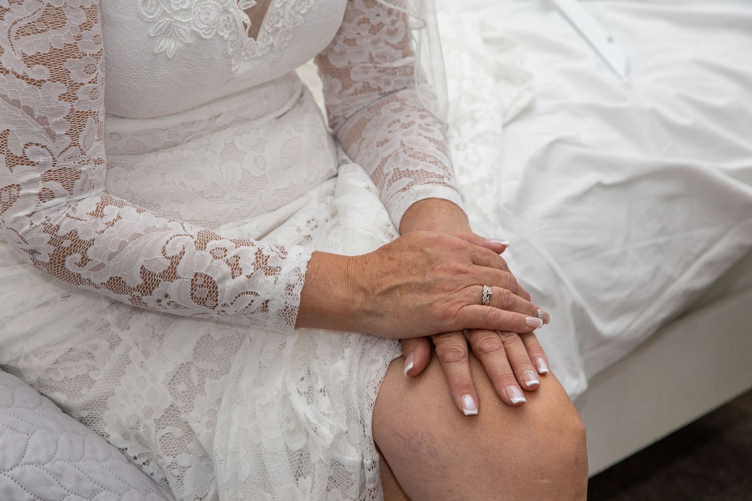 Bride in white lace wedding dress sitting with hands folded on knee, wearing engagement ring.