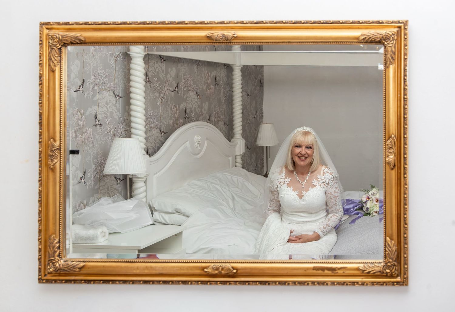Smiling bride in white lace dress and veil seated on bed, reflected in ornate gold-framed mirror in elegant bedroom.