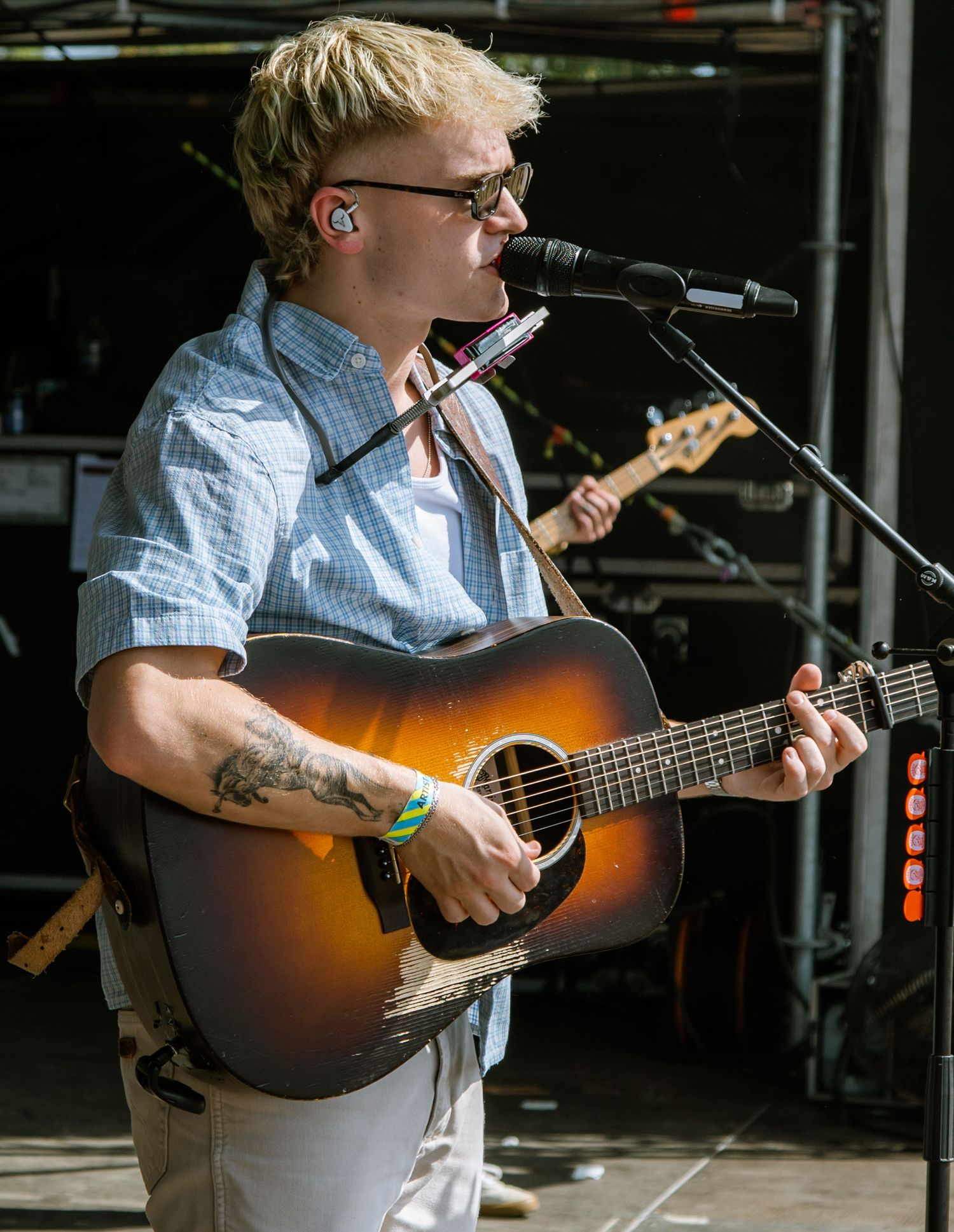 Blonde musician with tattoos plays acoustic guitar and sings into microphone on outdoor stage during live performance.