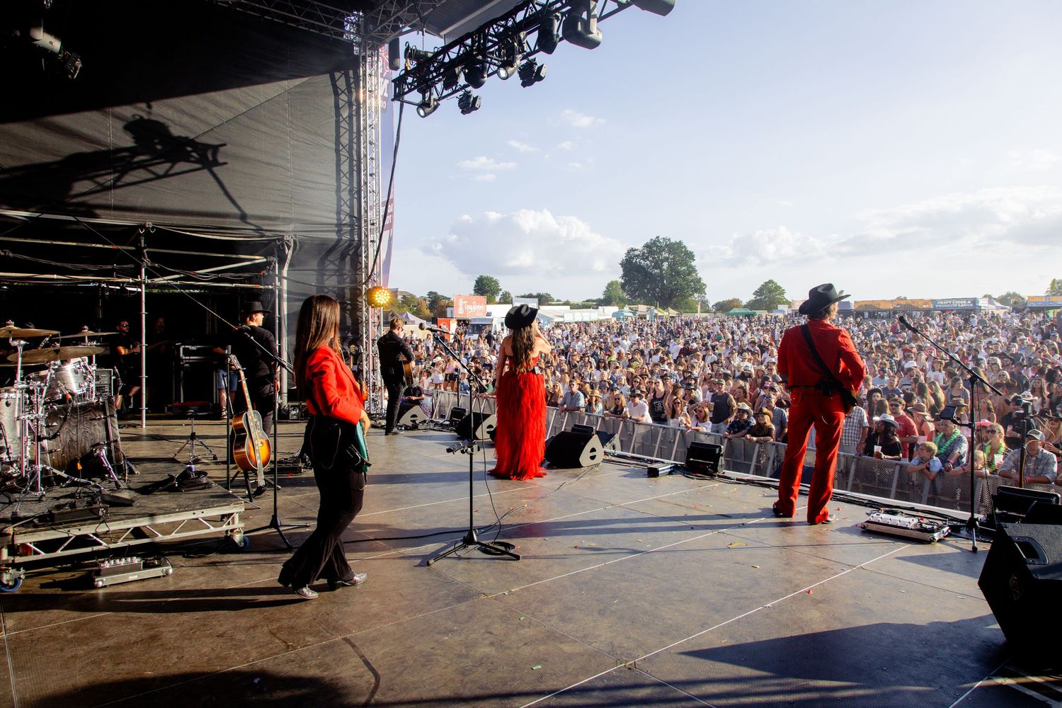 Band performing on outdoor stage to large festival crowd under blue sky.