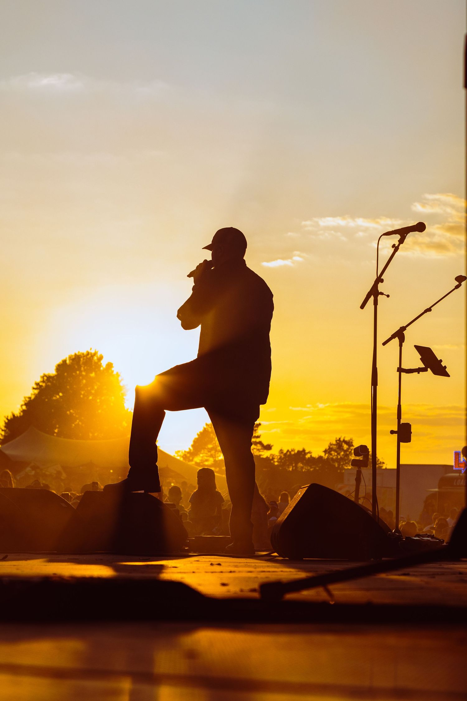 Silhouette of a performer singing on stage at sunset with microphone stands and crowd in golden backlight.