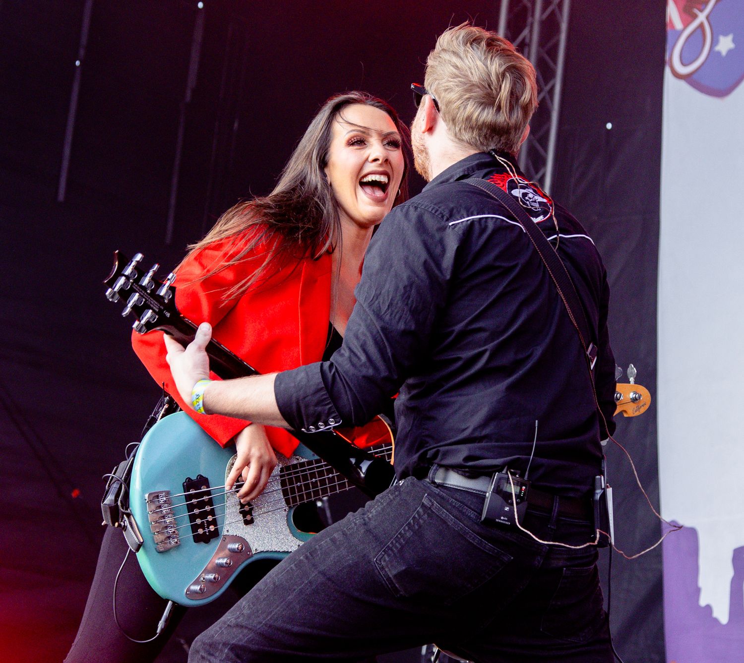 Two musicians share an energetic moment on stage, woman in red holds bass guitar while embracing male guitarist.