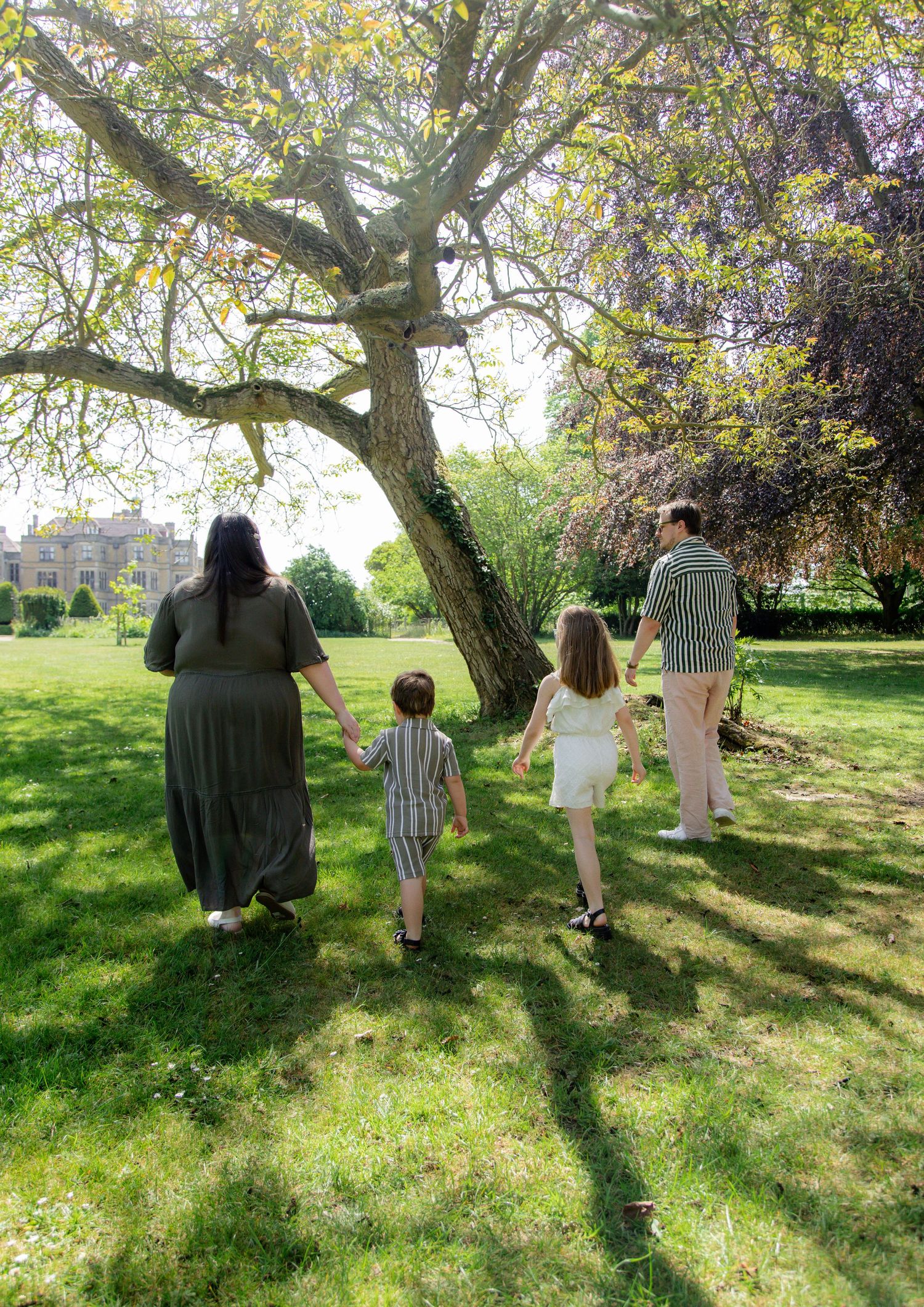 Family of four walking hand-in-hand through a lush green park beneath a large tree on a sunny day.