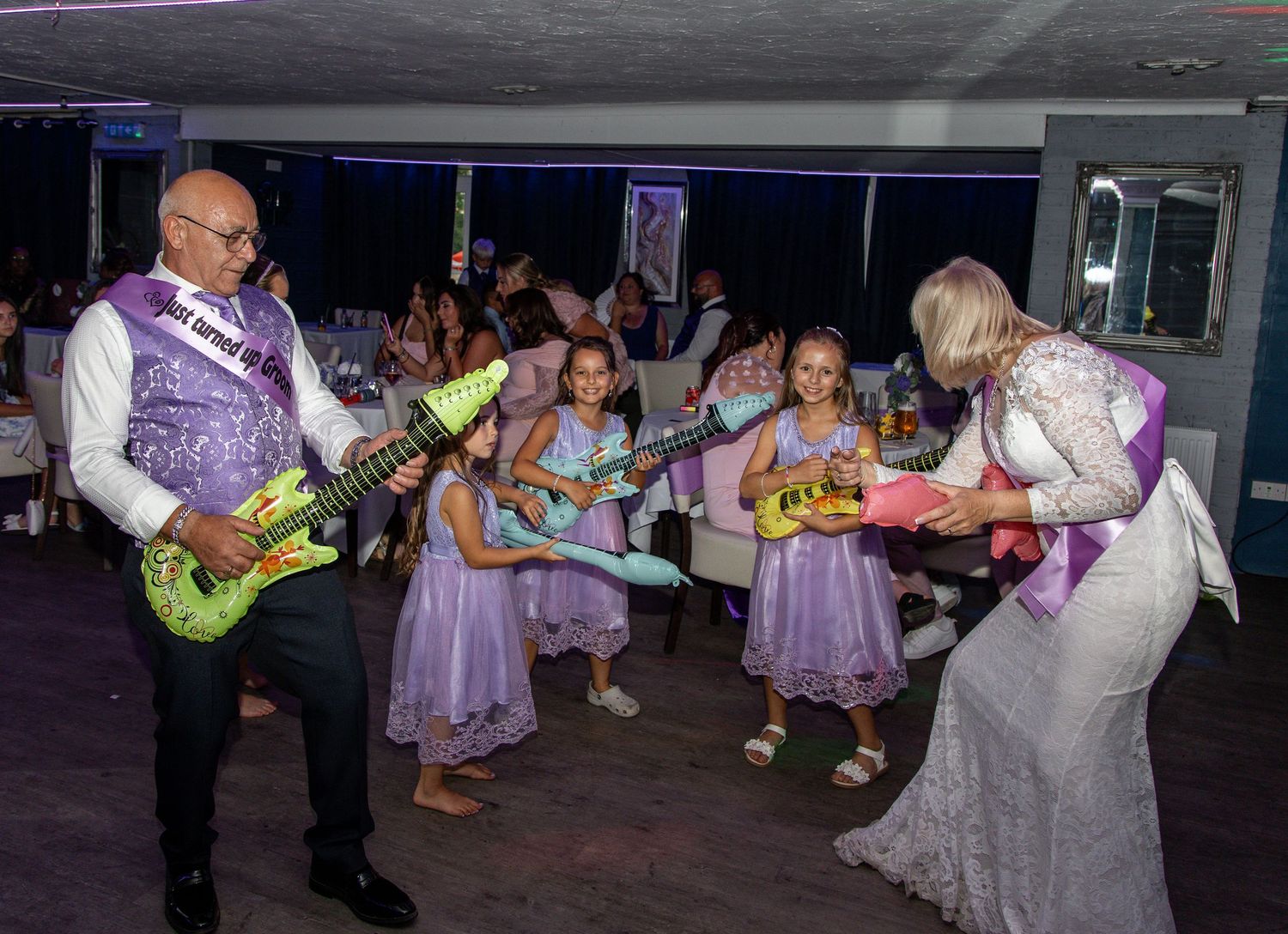 Bride and groom celebrate at wedding reception dancing with toy guitars alongside flower girls.