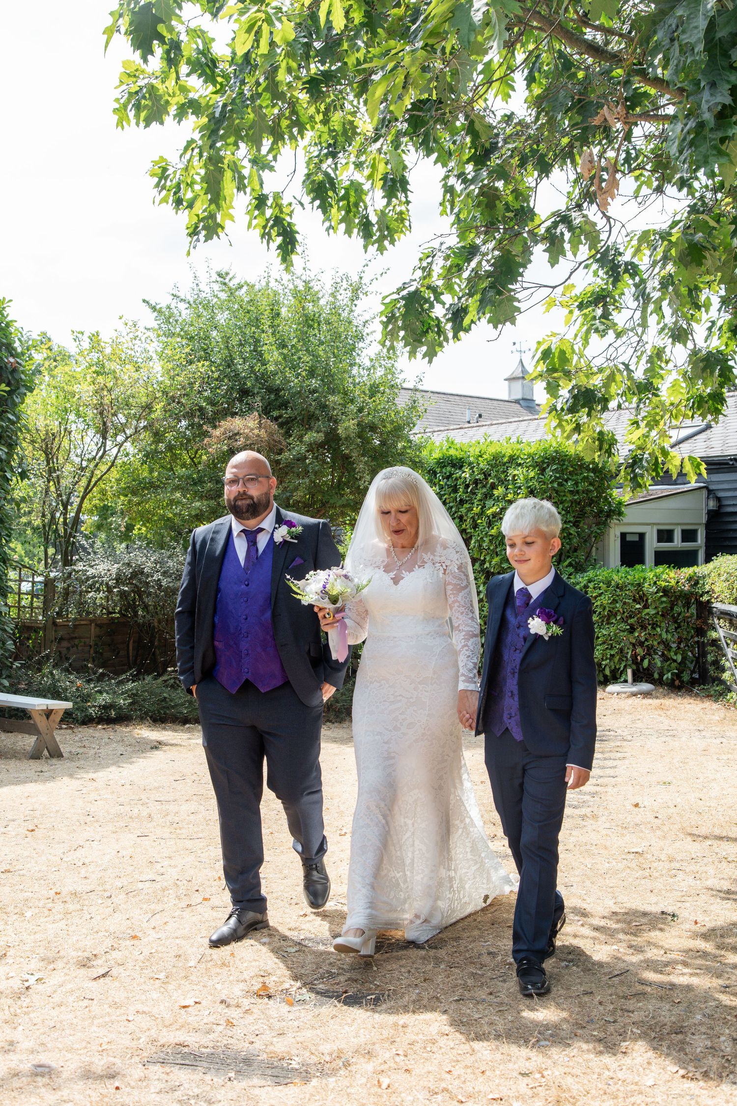 Bride in white lace gown walks with two groomsmen in purple vests through a sunny garden setting.