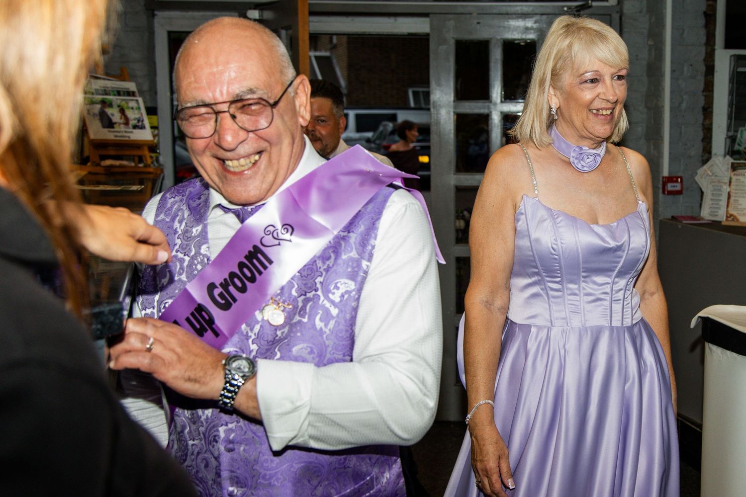 Elderly groom wearing purple sash smiles at wedding celebration with woman in lavender dress nearby.