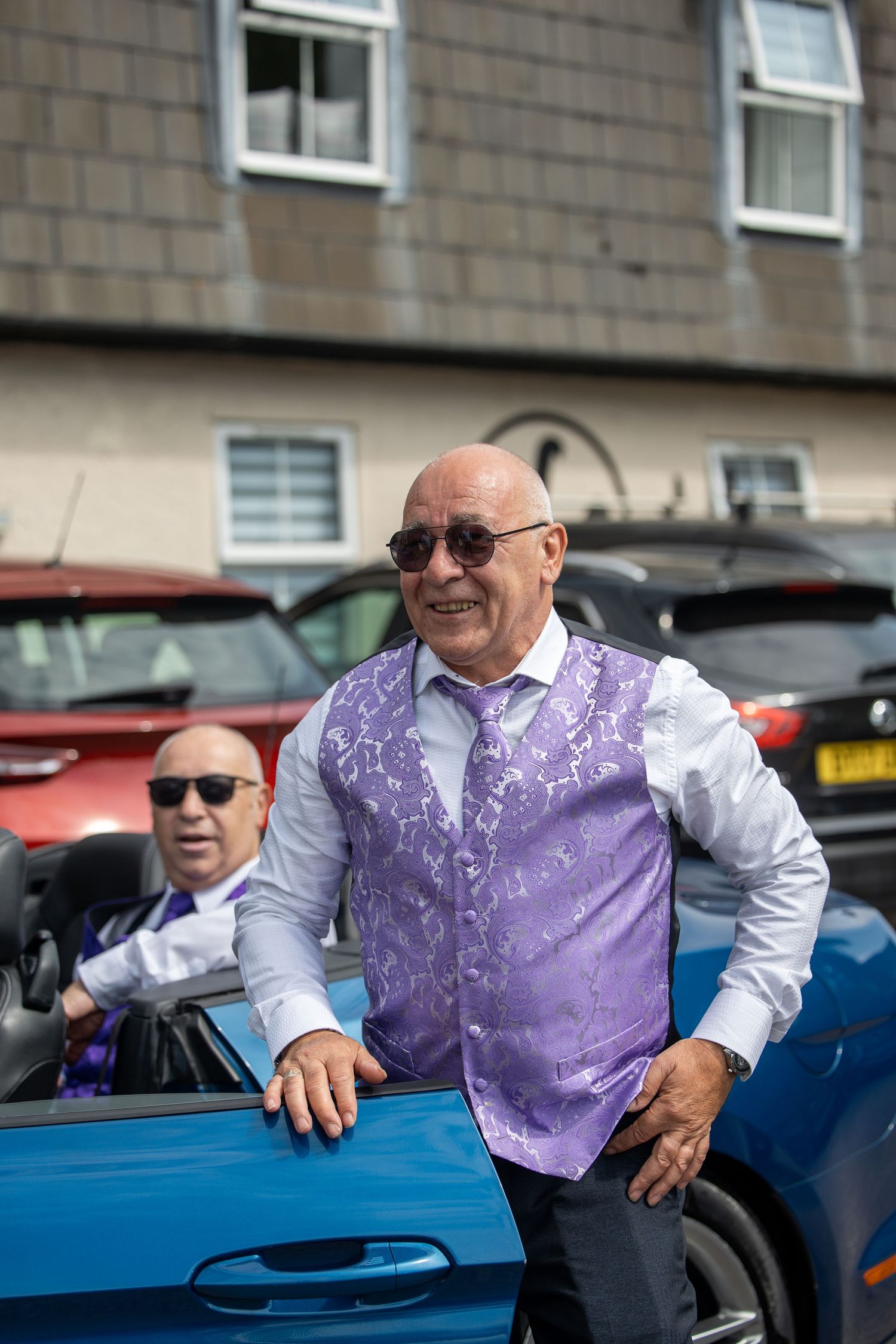 Smiling bald man in purple floral vest and sunglasses stands beside a blue convertible car outdoors.