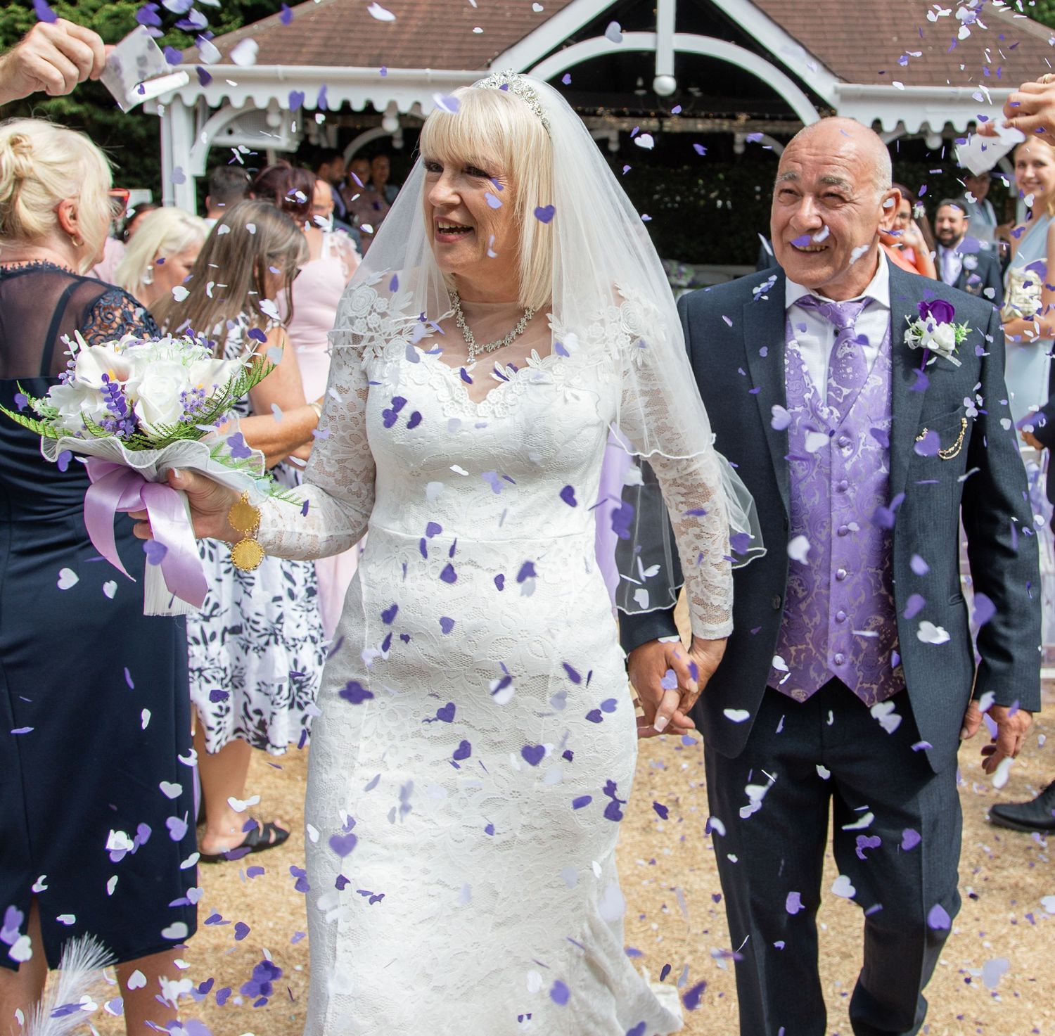 Elderly bride and groom walk through purple confetti shower after wedding ceremony outdoors.