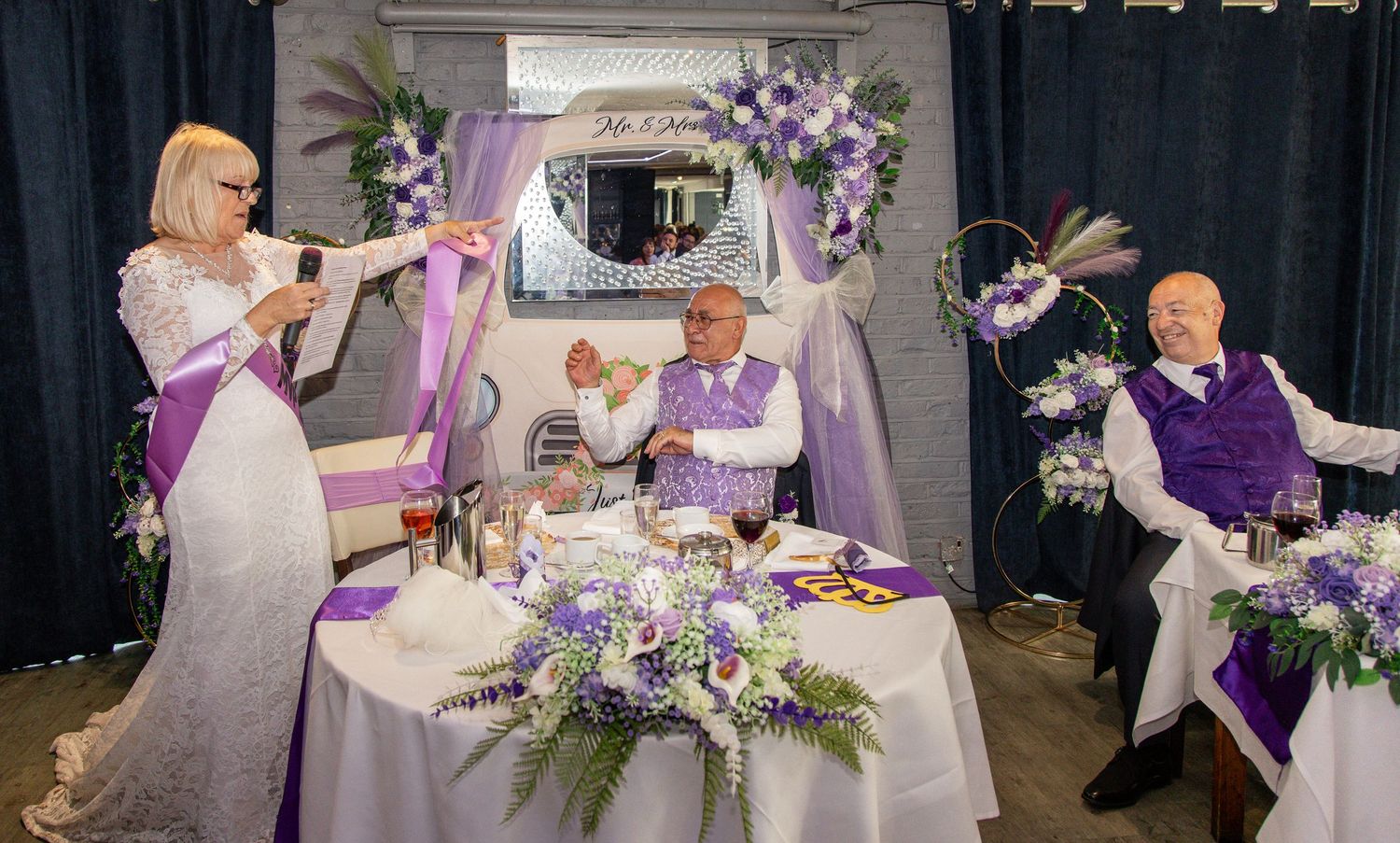 Elegant wedding reception with purple and white decor, floral arrangements, and guests in matching attire at a decorated table.
