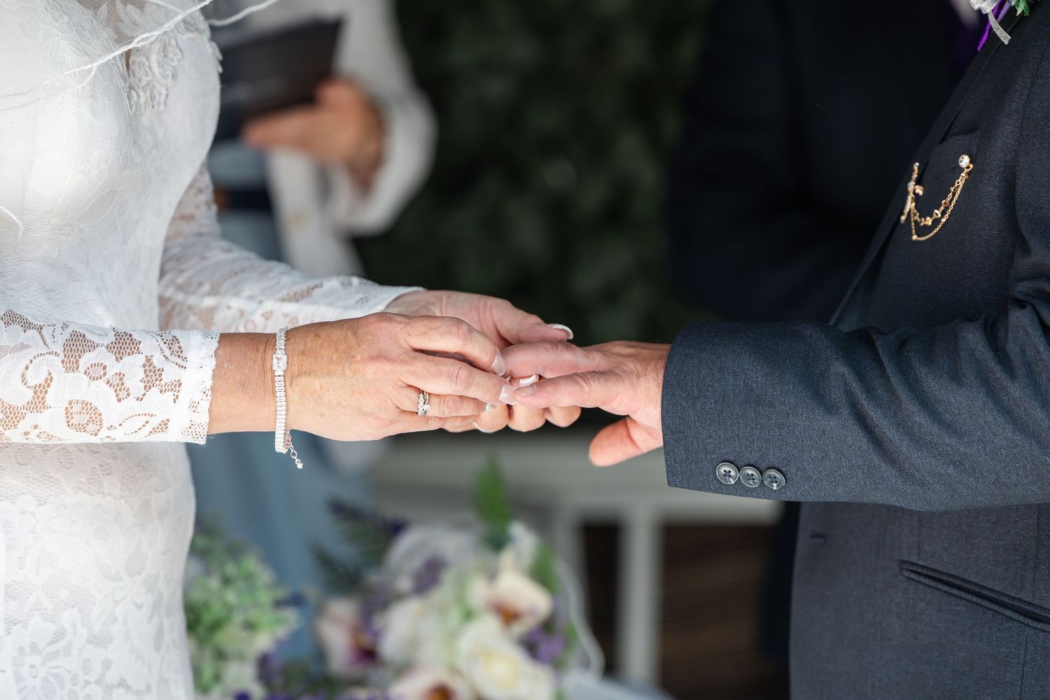 Bride placing a wedding ring on groom's finger during outdoor ceremony, wearing lace dress and bracelet.