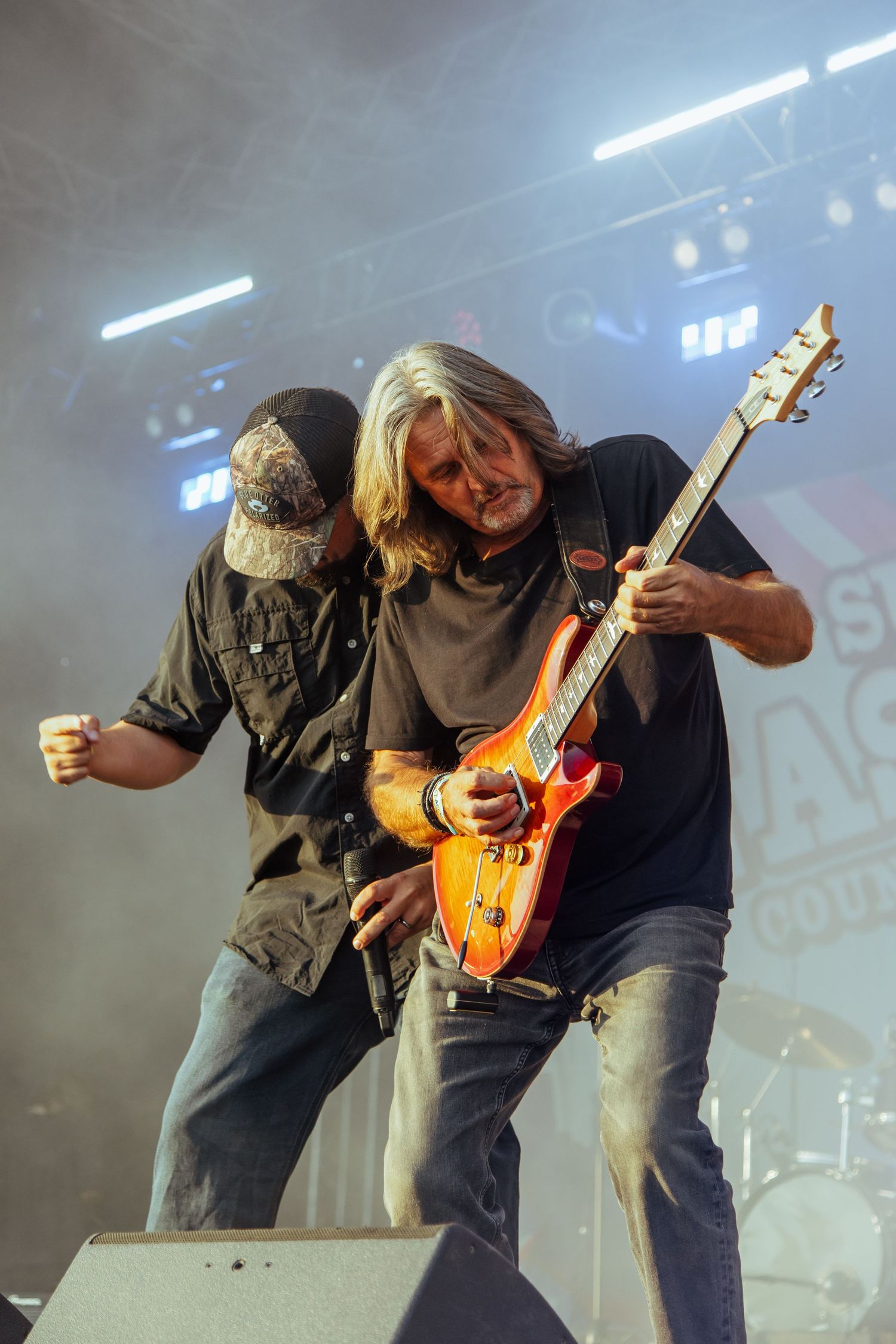 Two rock musicians performing on stage, one playing an orange electric guitar under dramatic stage lighting.