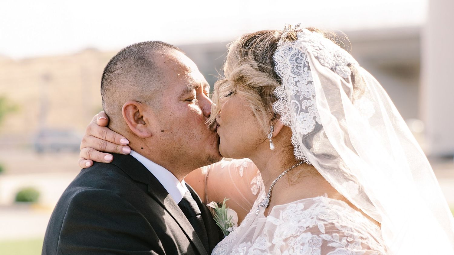 A bride and groom share a romantic kiss outdoors on their wedding day, with the bride wearing a lace veil.