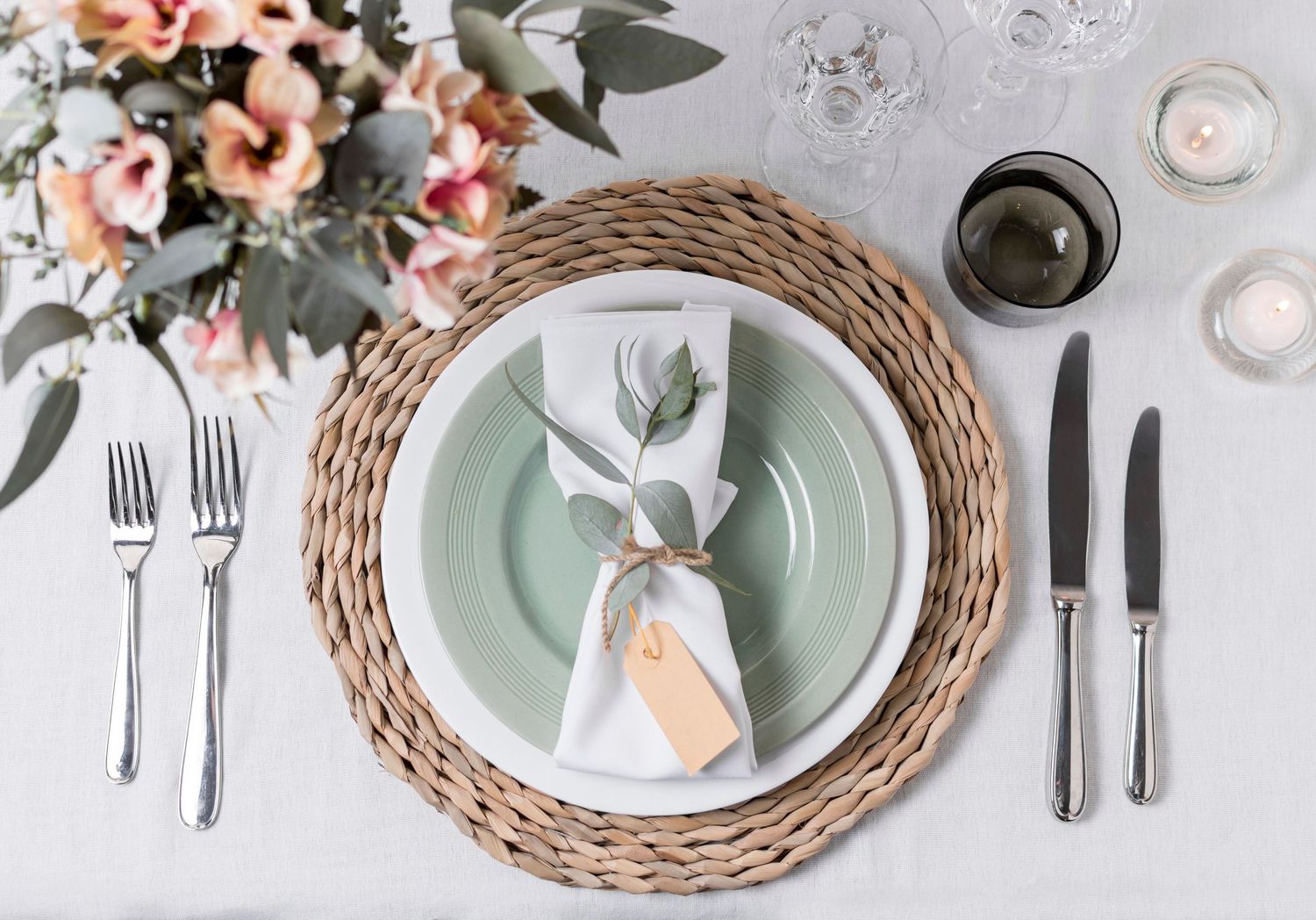 Elegant table setting with sage green plate on woven charger, white napkin, silverware, candles, and pink floral centerpiece.