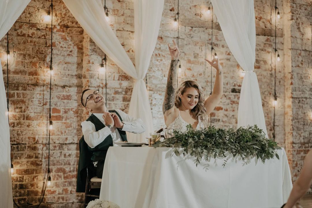 Bride and groom celebrating at wedding reception table with fairy lights and rustic brick wall backdrop.