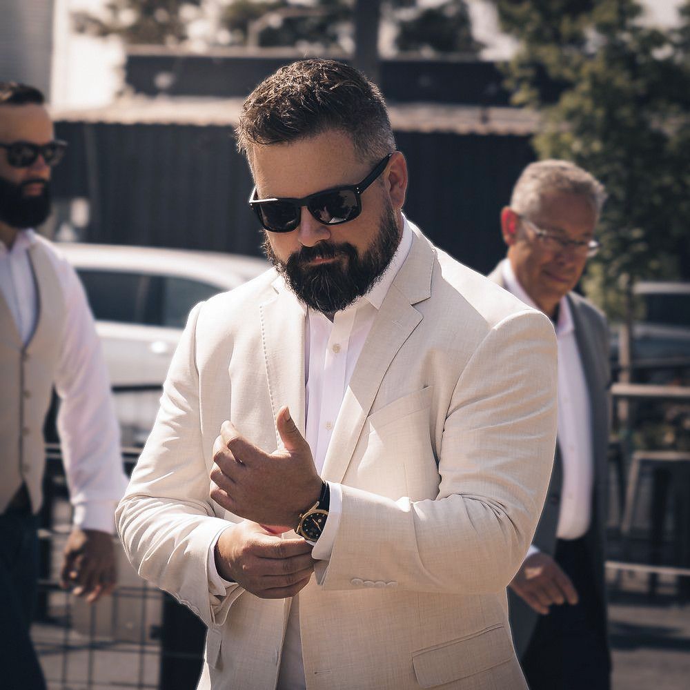 Bearded man in white suit adjusting cufflinks outdoors, wearing sunglasses and a gold watch at a formal event.