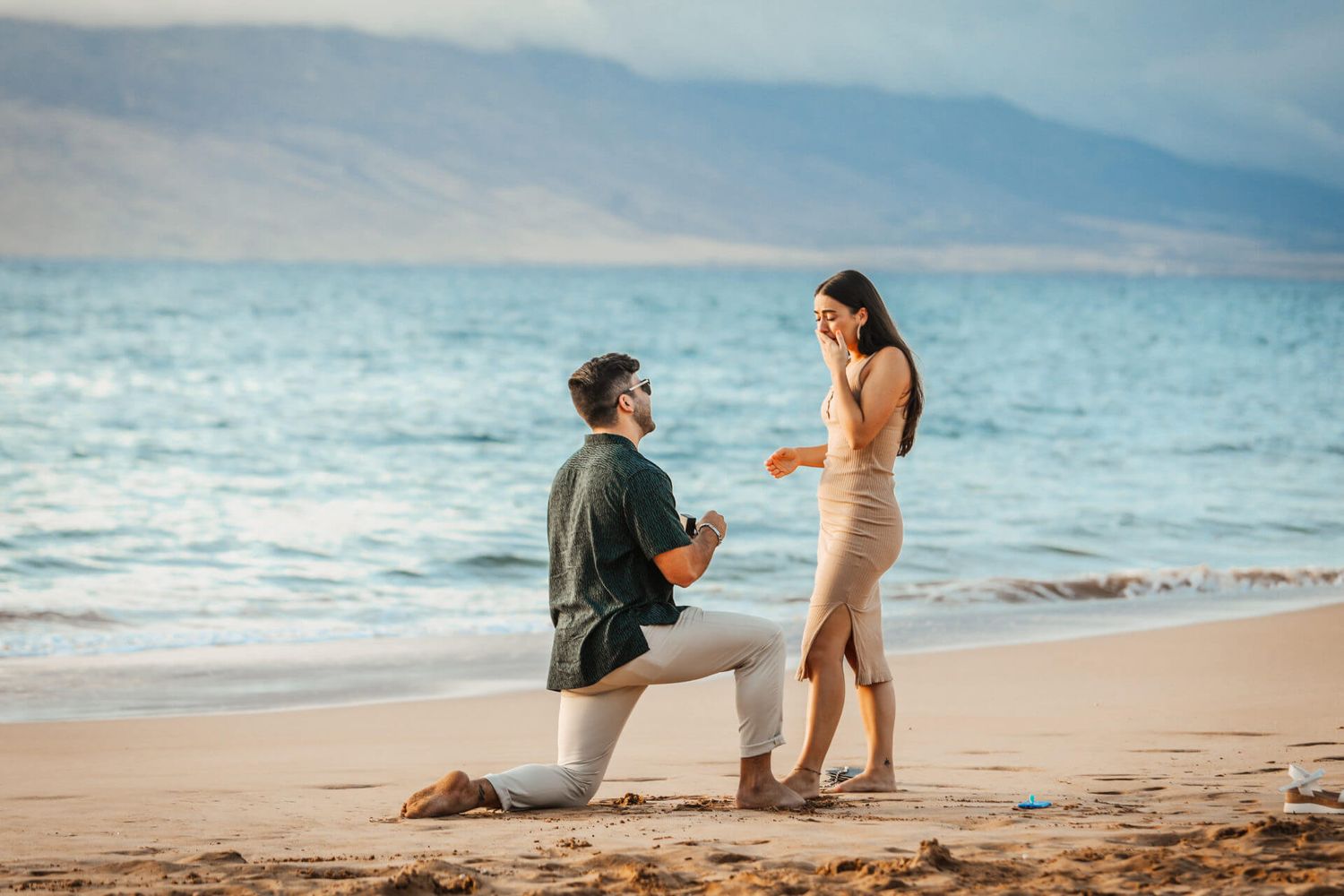 Man proposing to woman on a sandy beach with ocean and mountains in the background.