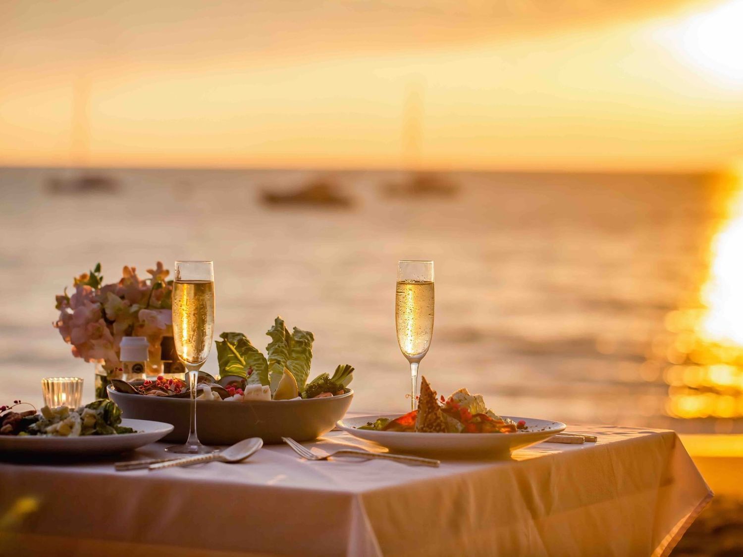 Romantic beachside dinner at sunset with champagne flutes, fresh salads, and seafood on a white tablecloth.