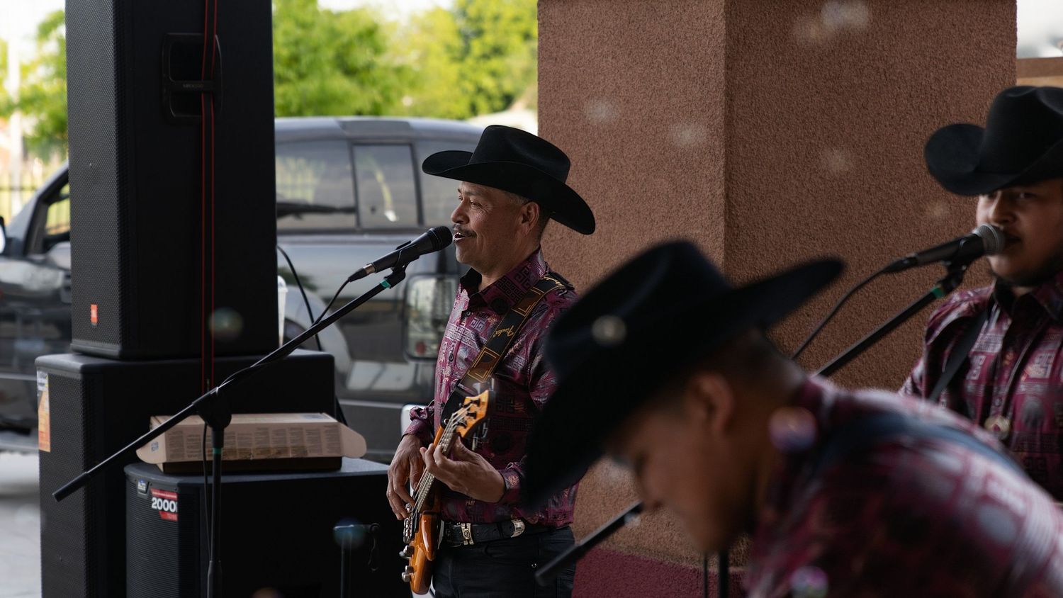 Three musicians in black cowboy hats perform country music outdoors, with guitarist playing at microphone stand.