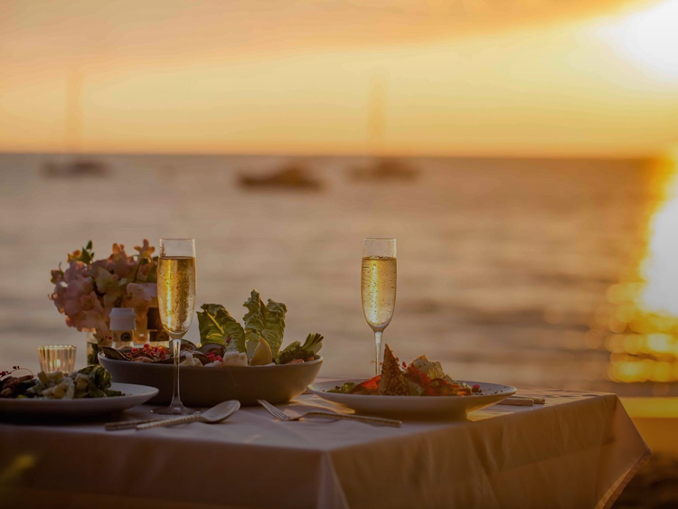 Romantic beachside dinner for two with champagne glasses and gourmet dishes set against a stunning golden ocean sunset.Merced Wedding Photog