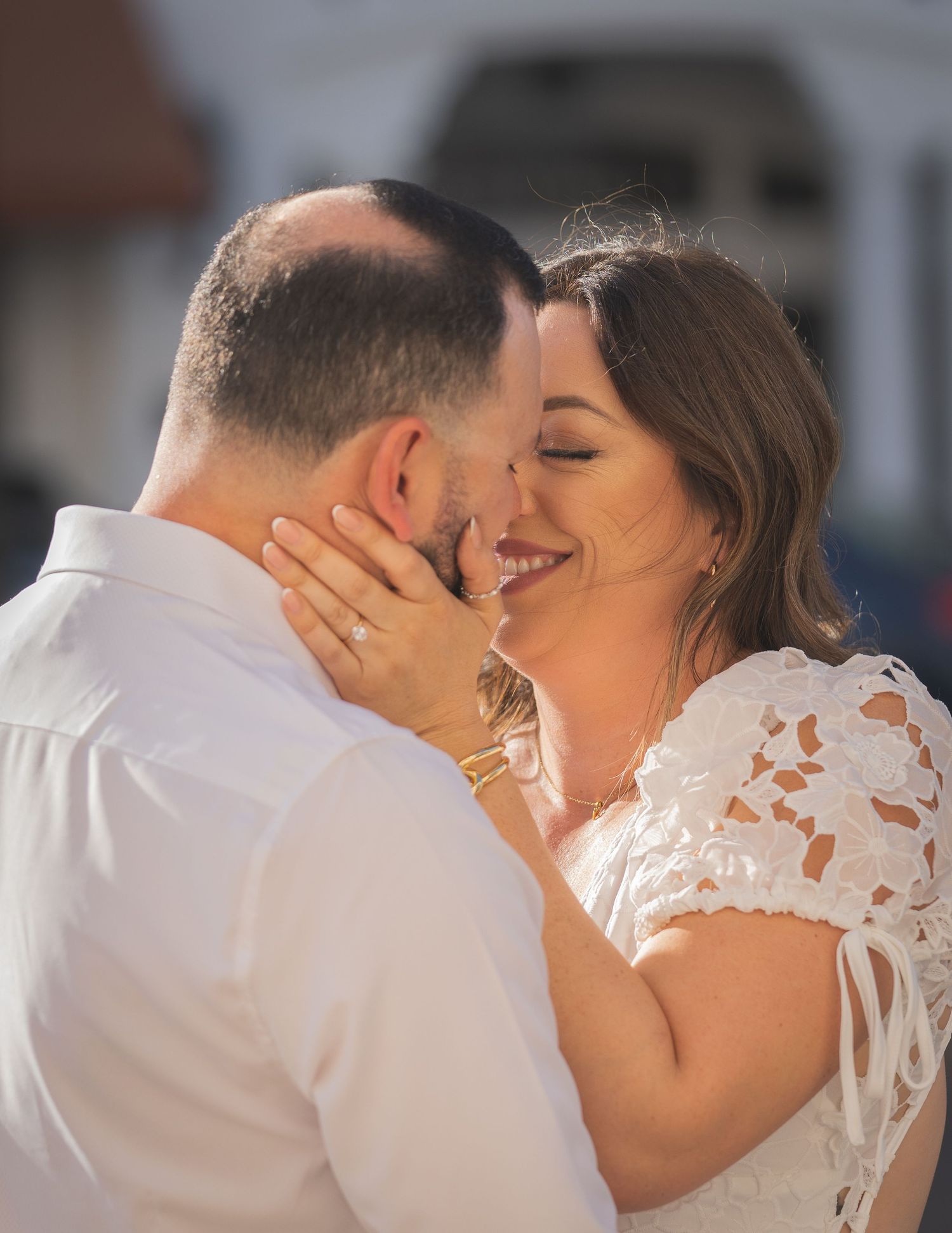 A couple shares a romantic almost-kiss moment, the smiling bride in white lace touching the grooms face outdoors.