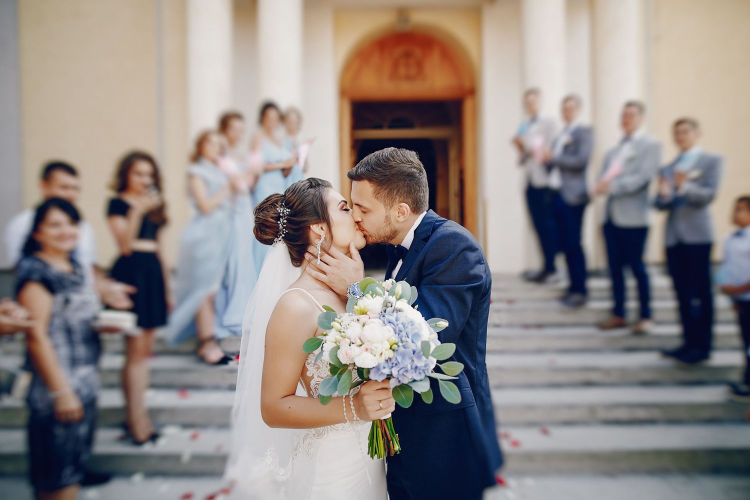 Newlyweds share a kiss outside a church, bride holding a white and purple bouquet, guests watching on the steps.