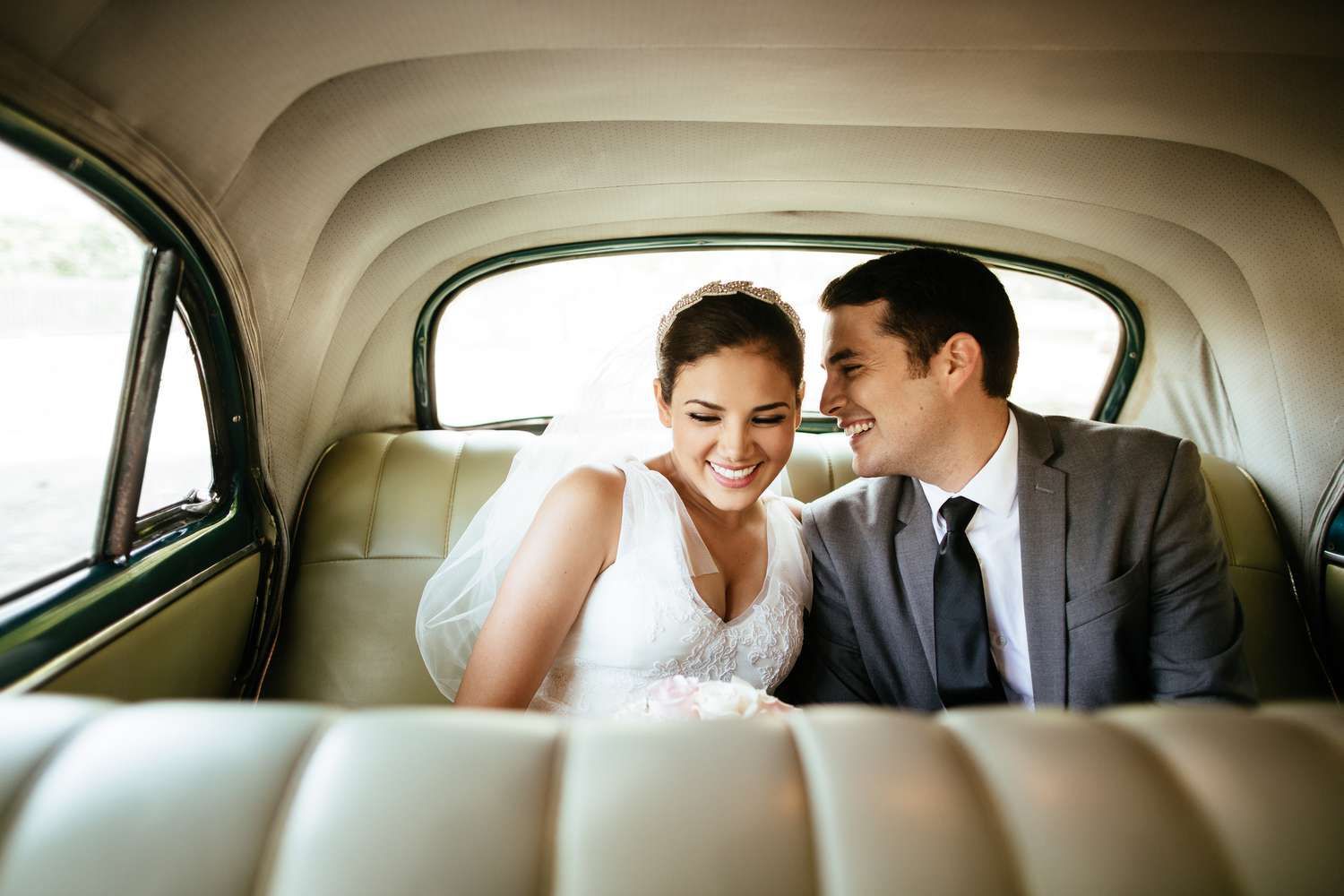 Newlywed couple smiling intimately in vintage car backseat, bride in white veil and groom in gray suit.