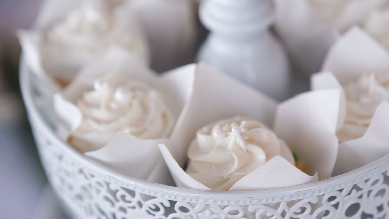 White frosted cupcakes in paper liners arranged in a decorative white lace tray with a salt shaker centerpiece.