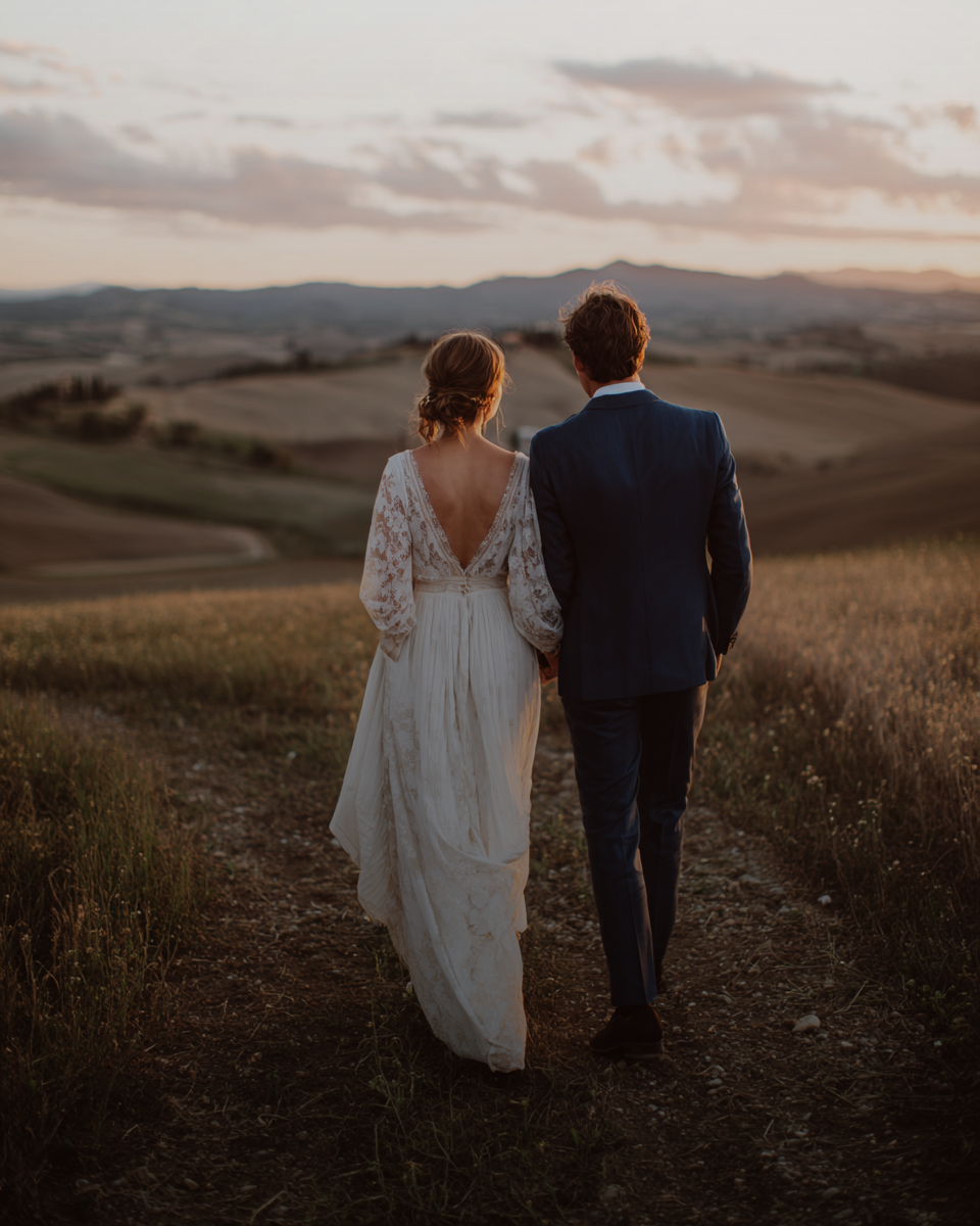 A bride and groom walk hand in hand through golden Tuscan hills at sunset, her lace gown trailing behind. Merced Wedding Photographer