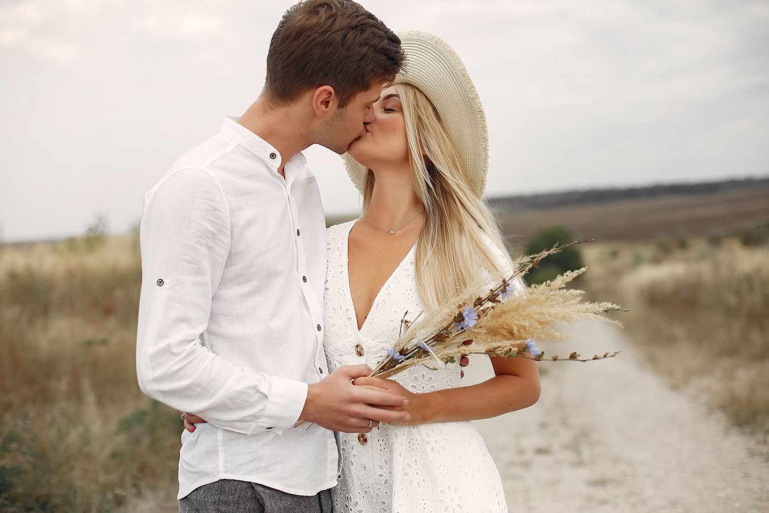 A romantic couple kissing outdoors, woman in white dress and hat holding wildflowers bouquet in a scenic field.