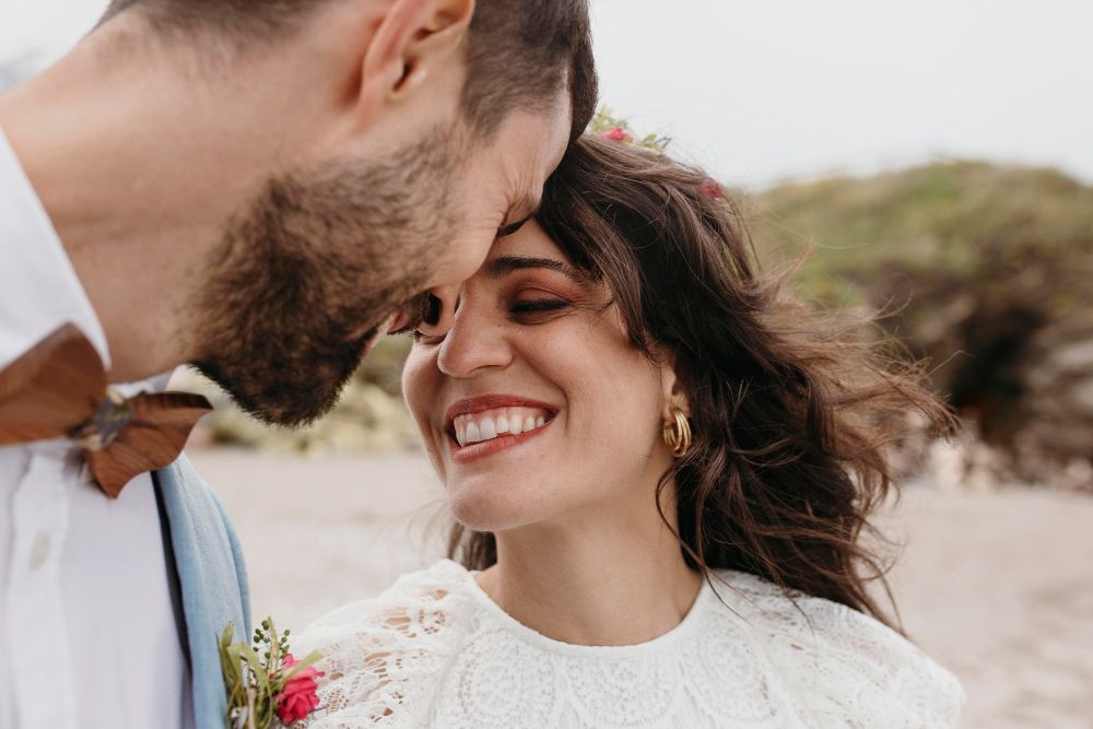 A smiling bride receives a kiss on the forehead from her groom outdoors on a beach wedding day. Merced Wedding Photographer