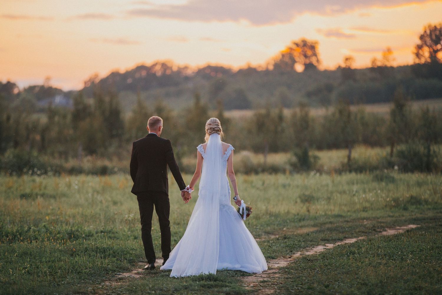 A couple in wedding attire walks along a scenic country path at sunset with golden light filtering through trees.