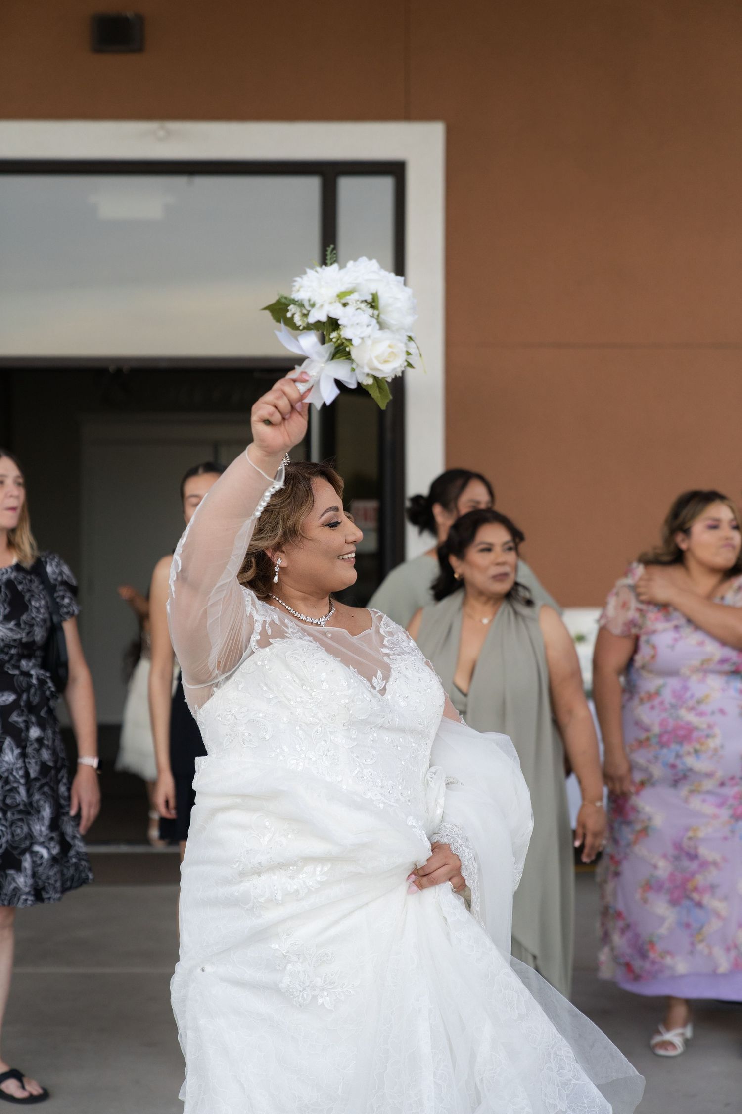 Bride in white lace gown tossing bouquet of white flowers at wedding reception with guests in background.