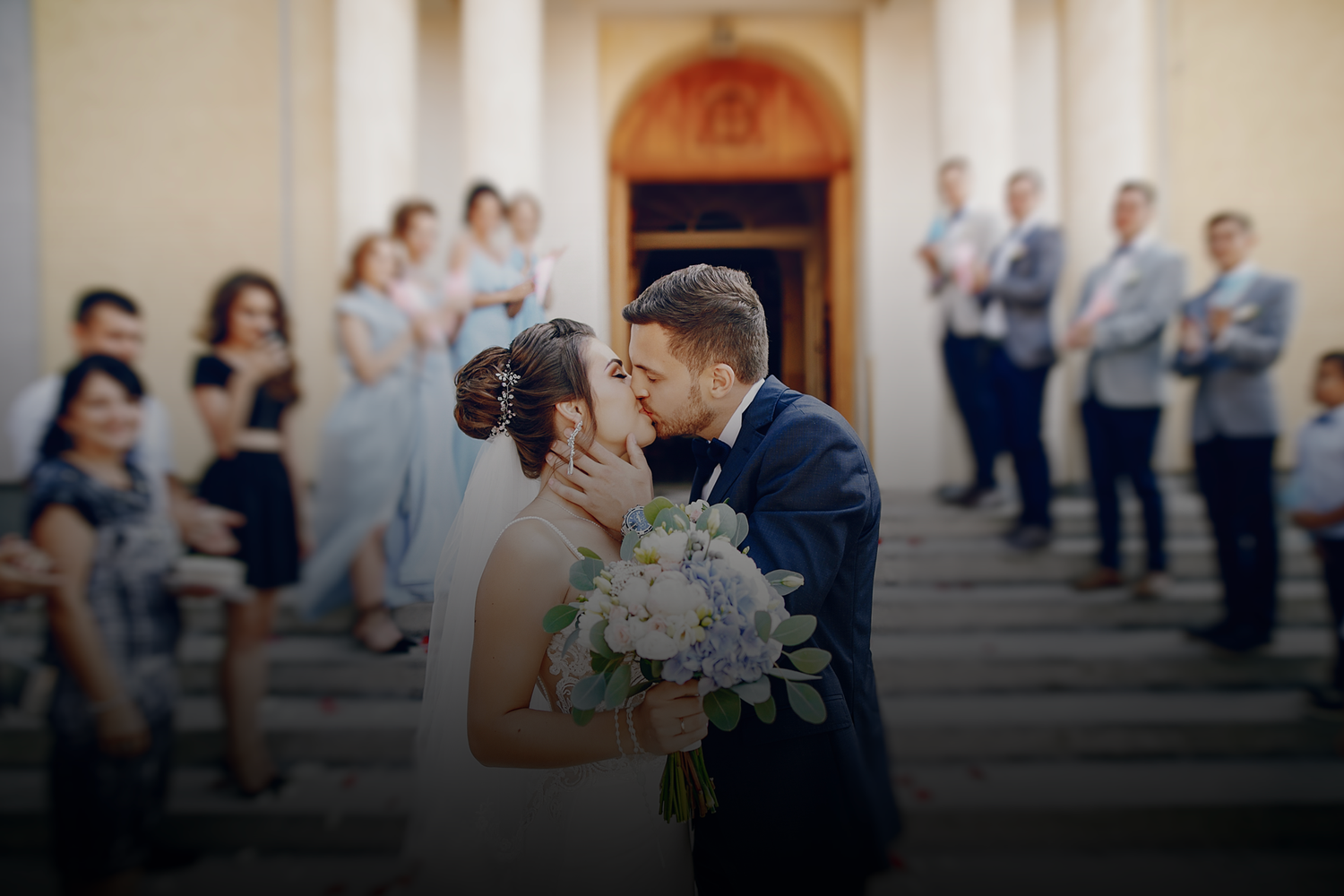 Bride and groom share a romantic kiss on church steps surrounded by wedding party guests in formal attire. Merced Wedding Photographer