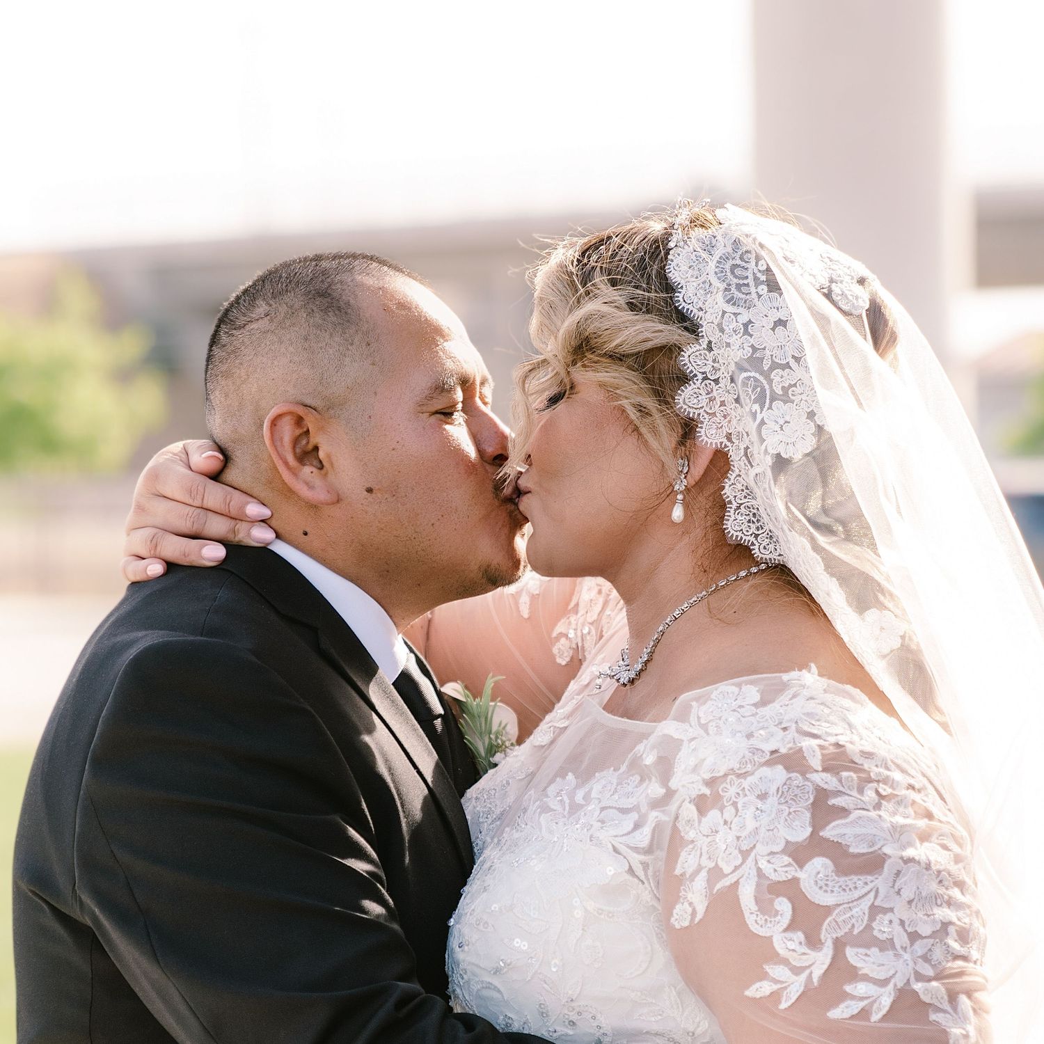 A bride and groom share a romantic kiss outdoors, bride wearing lace wedding dress and veil, groom in black suit.