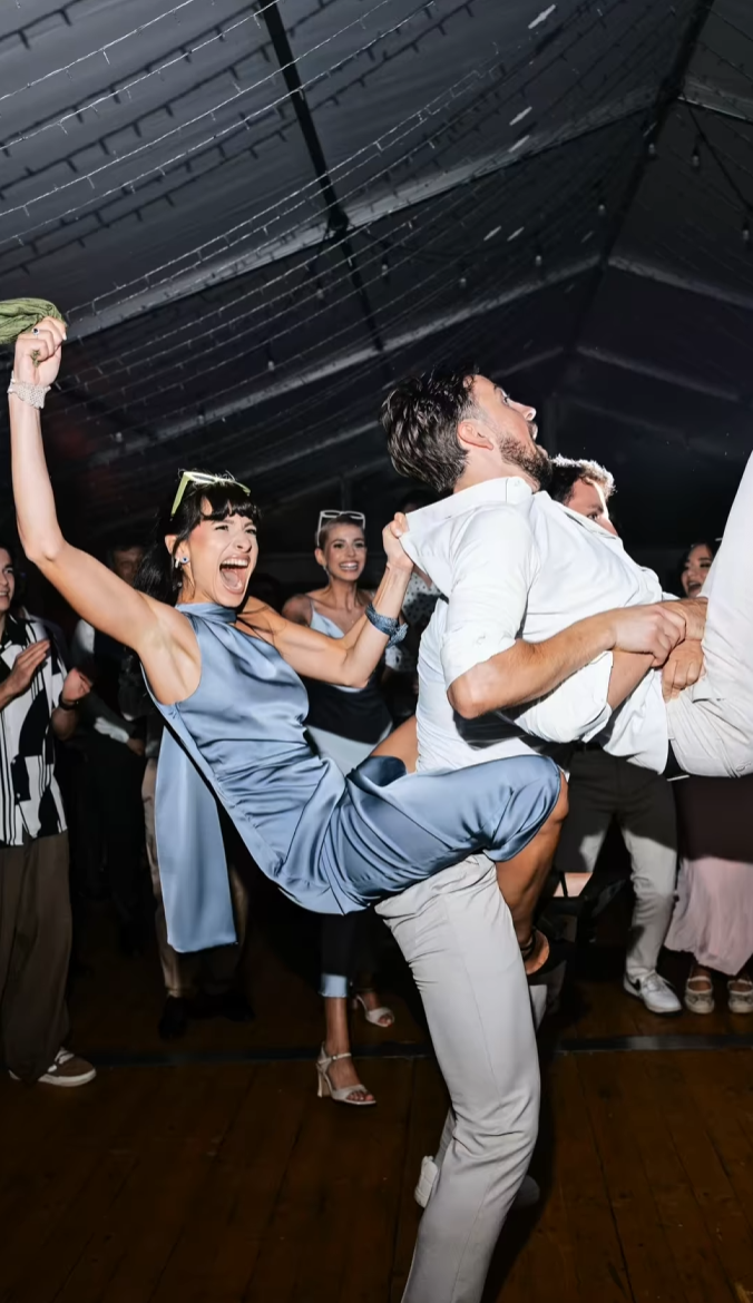 Couple dancing energetically at a wedding reception, woman in blue dress lifted by man in white shirt.